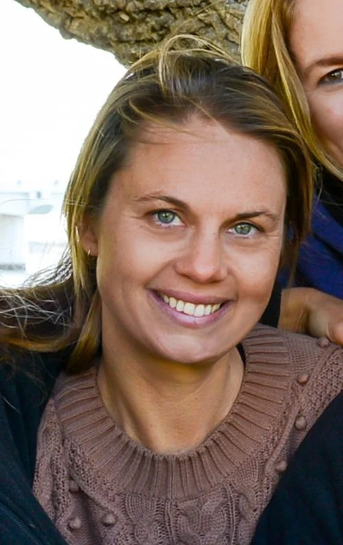 A woman with blonde hair, blue eyes, and a bright smile, wearing a brown knitted sweater. Part of another person's face is visible on her right side.