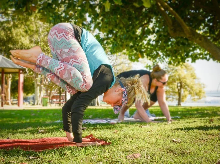 Two women and a girl practicing yoga outdoors on a sunny day in a park with trees and grass, in a downward dog pose.