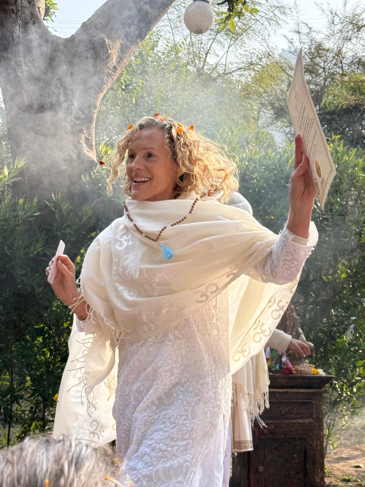 A woman with curly blonde hair wearing a white dress and shawl, holding a piece of paper, smiling joyfully outdoors beneath a tree with sunlight filtering through.