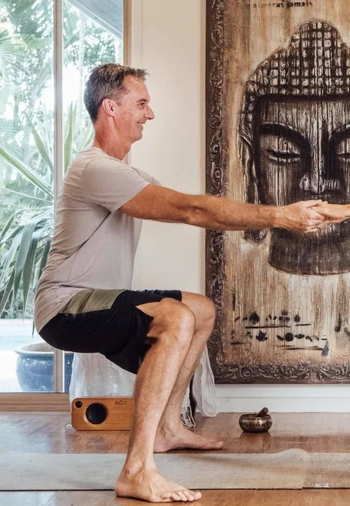 A man practicing yoga indoors, balancing on his toes with arms extended forward, in front of a large Buddha face painting on the wall.