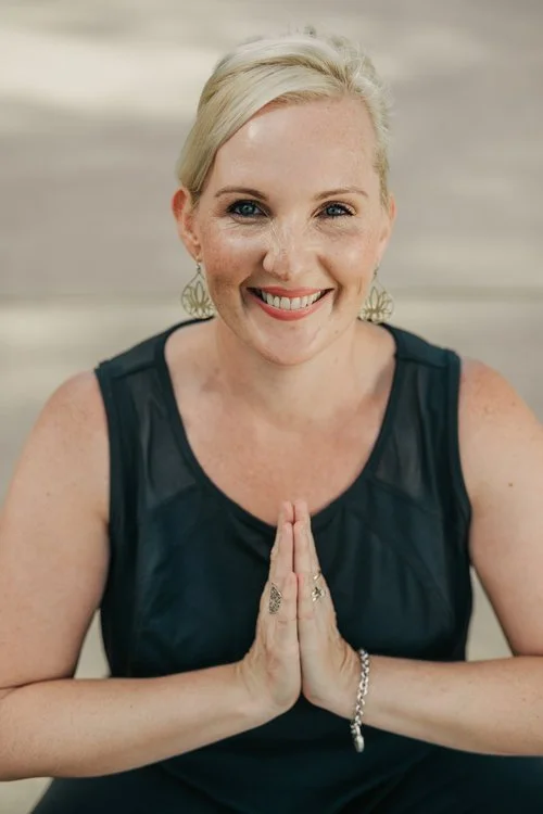 A woman with blonde hair smiling, holding her hands in a prayer position, wearing a black sleeveless top and earrings.
