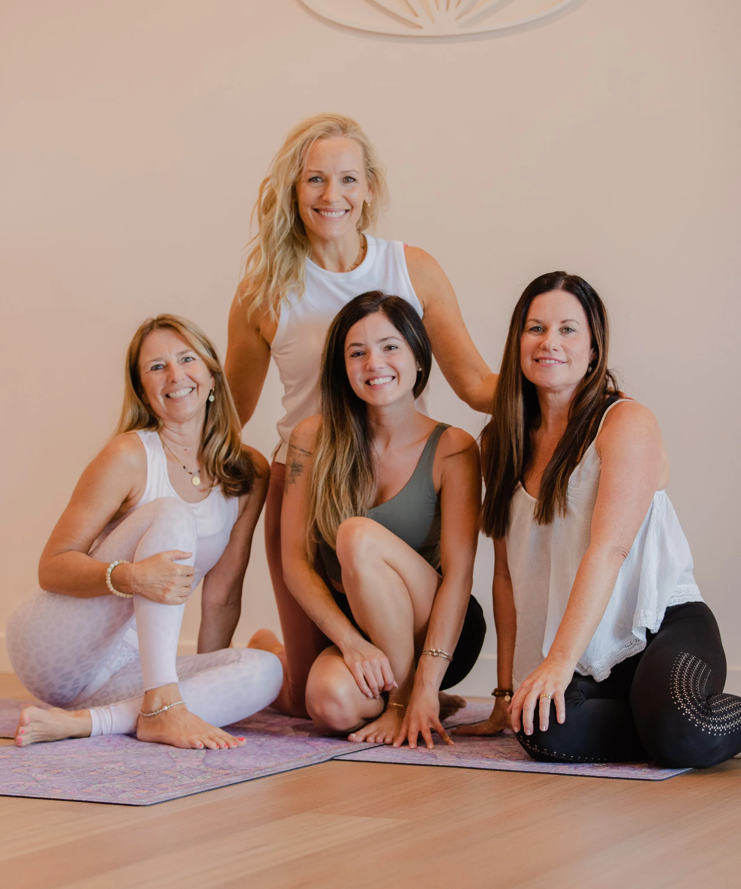 Five women in casual workout clothing posing together on yoga mats in a well-lit room.
