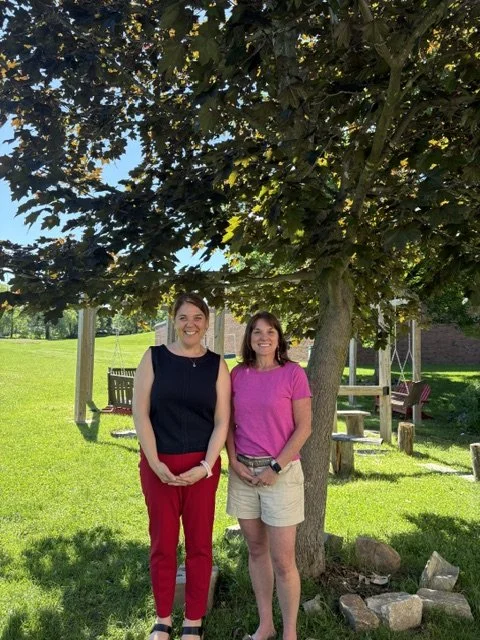 Two women standing under a large tree in a park, smiling at the camera. One woman is wearing a black top and red pants, the other is wearing a pink shirt and beige shorts.
