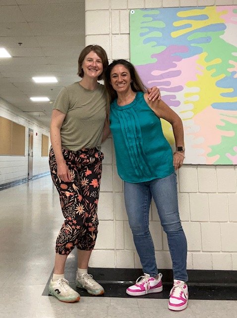 Two women smiling and posing together in a school hallway, standing beside a colorful abstract painting on the wall.