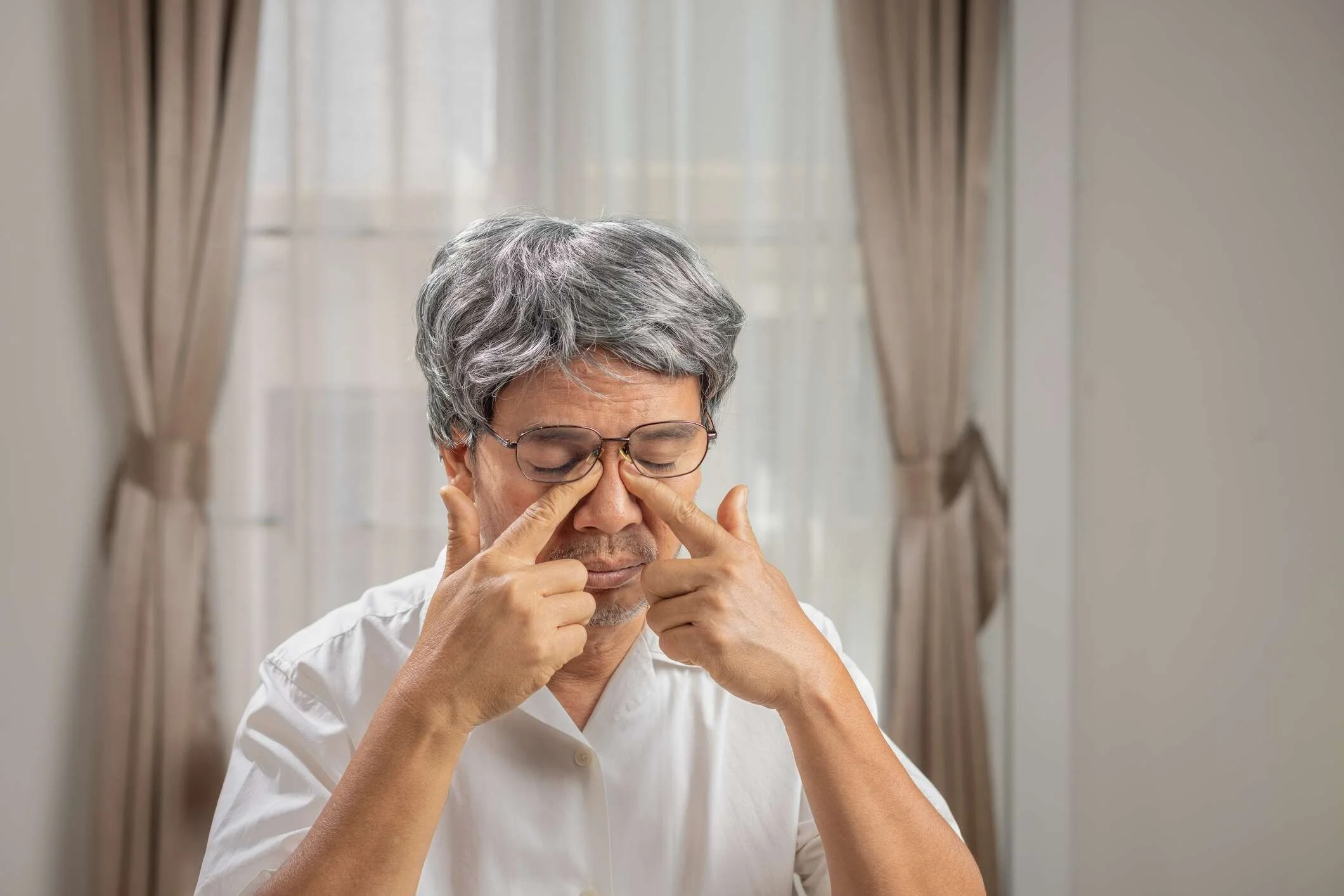 A senior man wearing a white shirt and glasses rubs his eyes.
