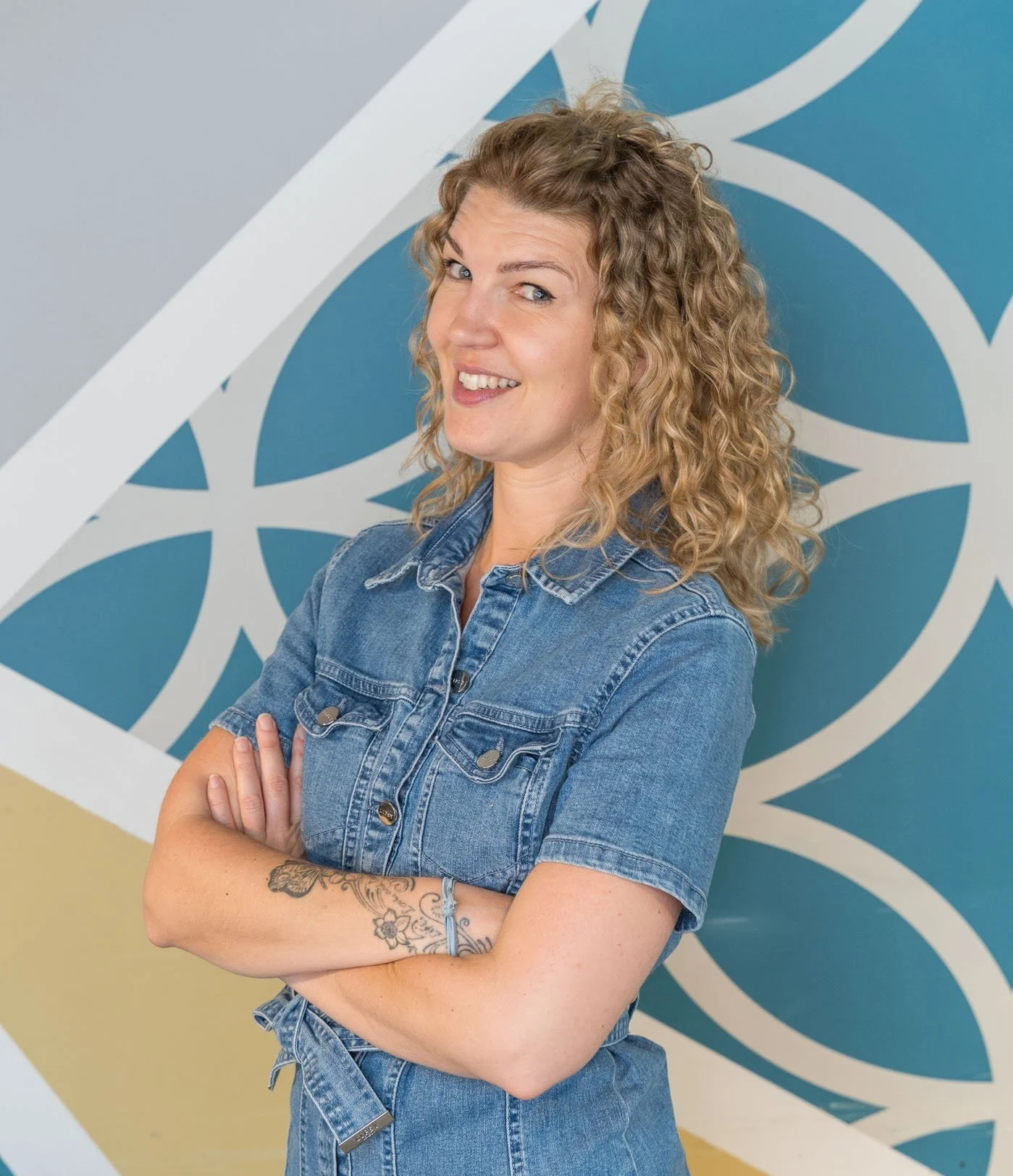 A woman with curly blonde hair, wearing a denim shirt and matching jeans, standing with arms crossed and smiling, in front of a blue and white patterned wall.