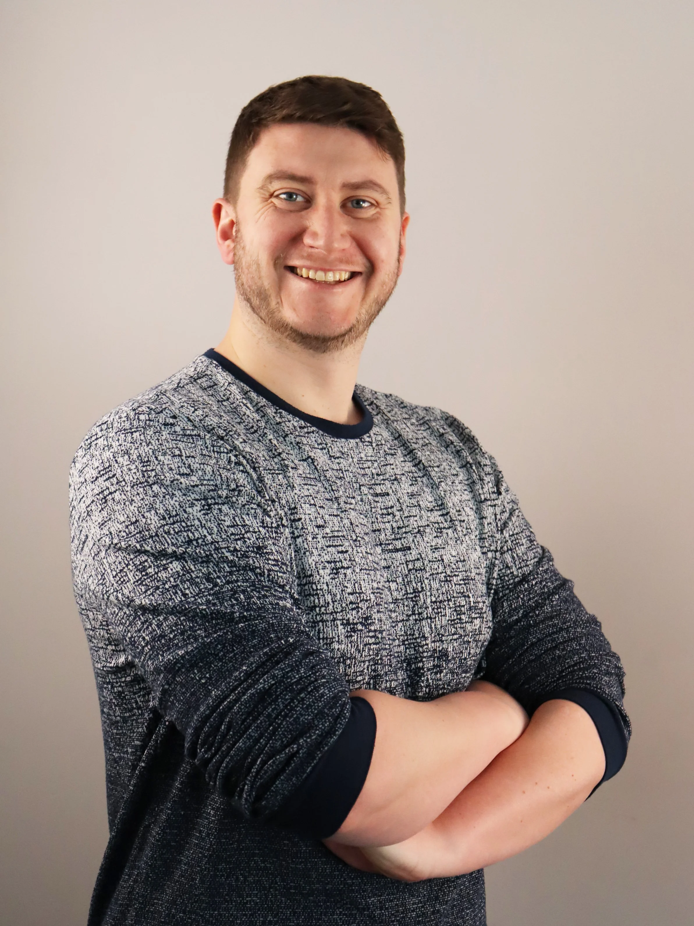 A smiling man with crossed arms, short brown hair, and a trimmed beard, wearing a patterned long-sleeve shirt, standing against a plain light-colored background.