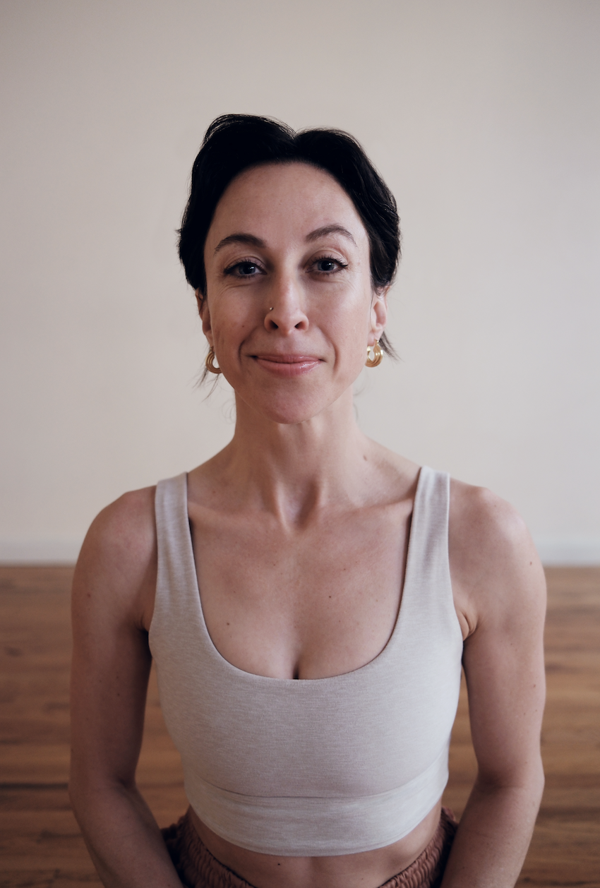 A woman practicing yoga on a pink yoga mat indoors, performing the cobra pose with her arms extended and chest lifted, in a living room with a brown couch and shelves on the wall.