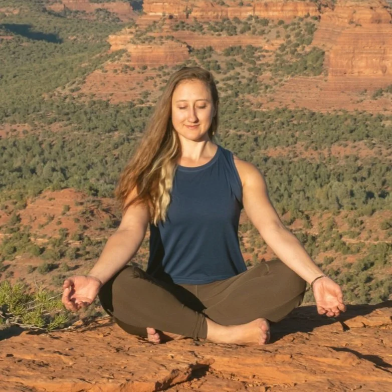 A woman meditating in a seated lotus position on a rocky ledge with a canyon in the background during sunset.