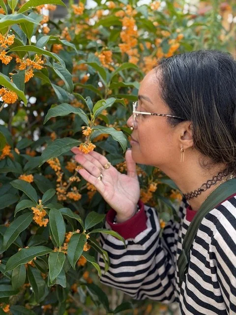 A woman with curly hair and glasses takes a selfie while doing yoga in a room filled with potted plants. She is on a green yoga mat, extending her arm with three fingers raised.