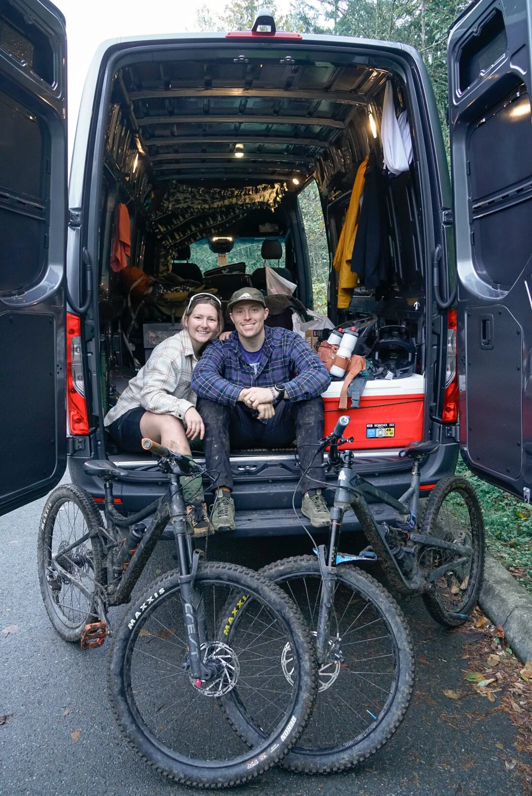 A couple sitting in the back of a van with mountain bikes in front.