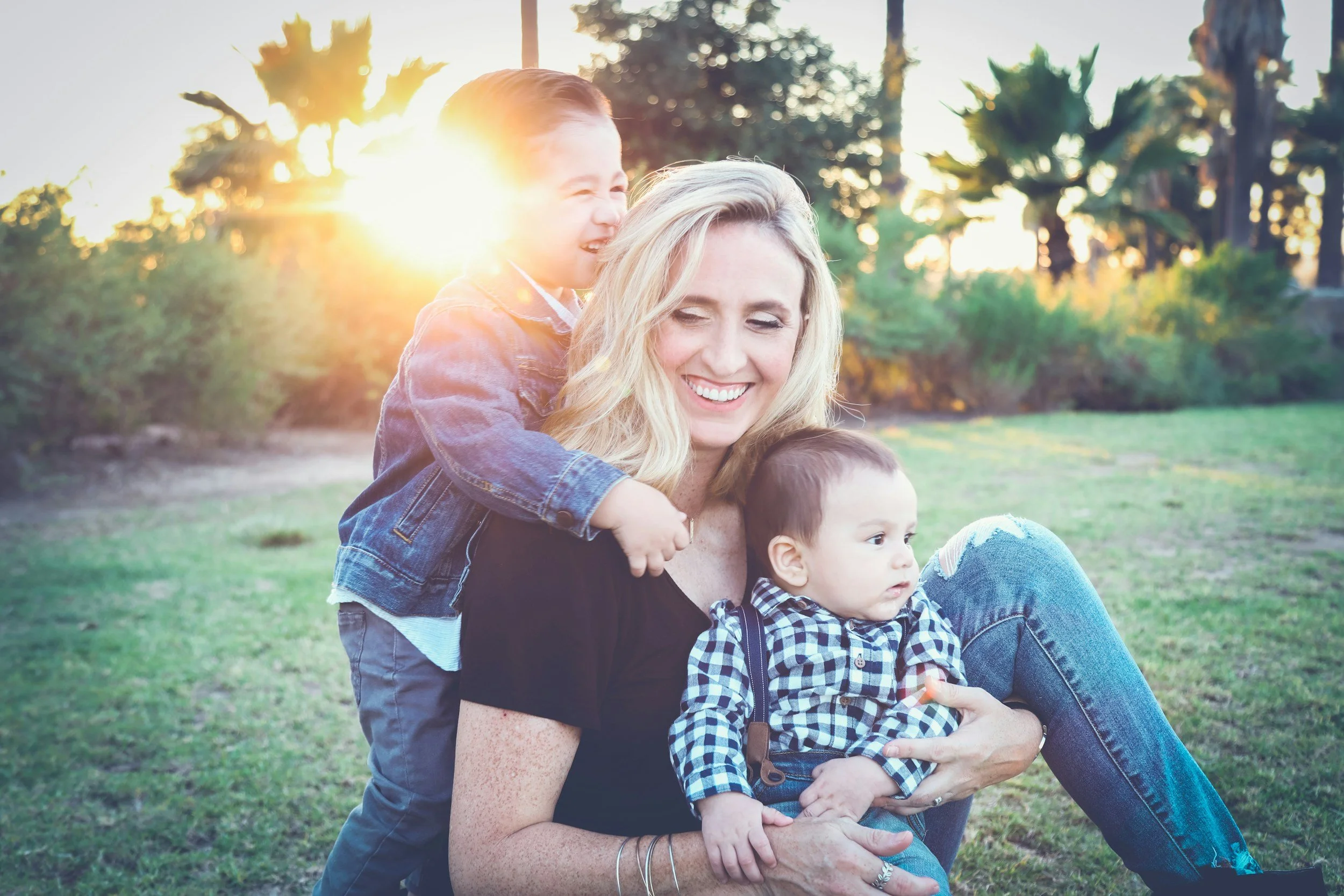 a nanny sits on the ground in a park in Indian Hill Ohio holding a little boy while a toddler hugs her and they laugh.