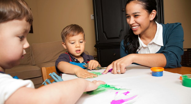 A nanny sits at a table with 2 little boys and they are painting hand prints on a large paper.