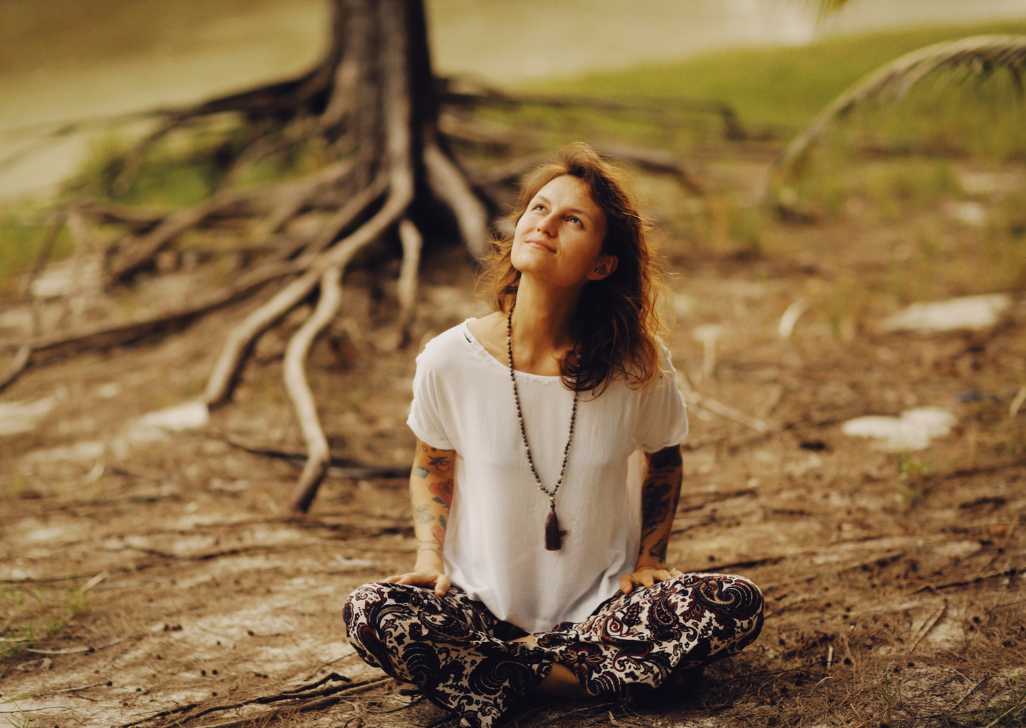 a woman sits cross legged in a forest. There is a tree with large visible roots behind her.