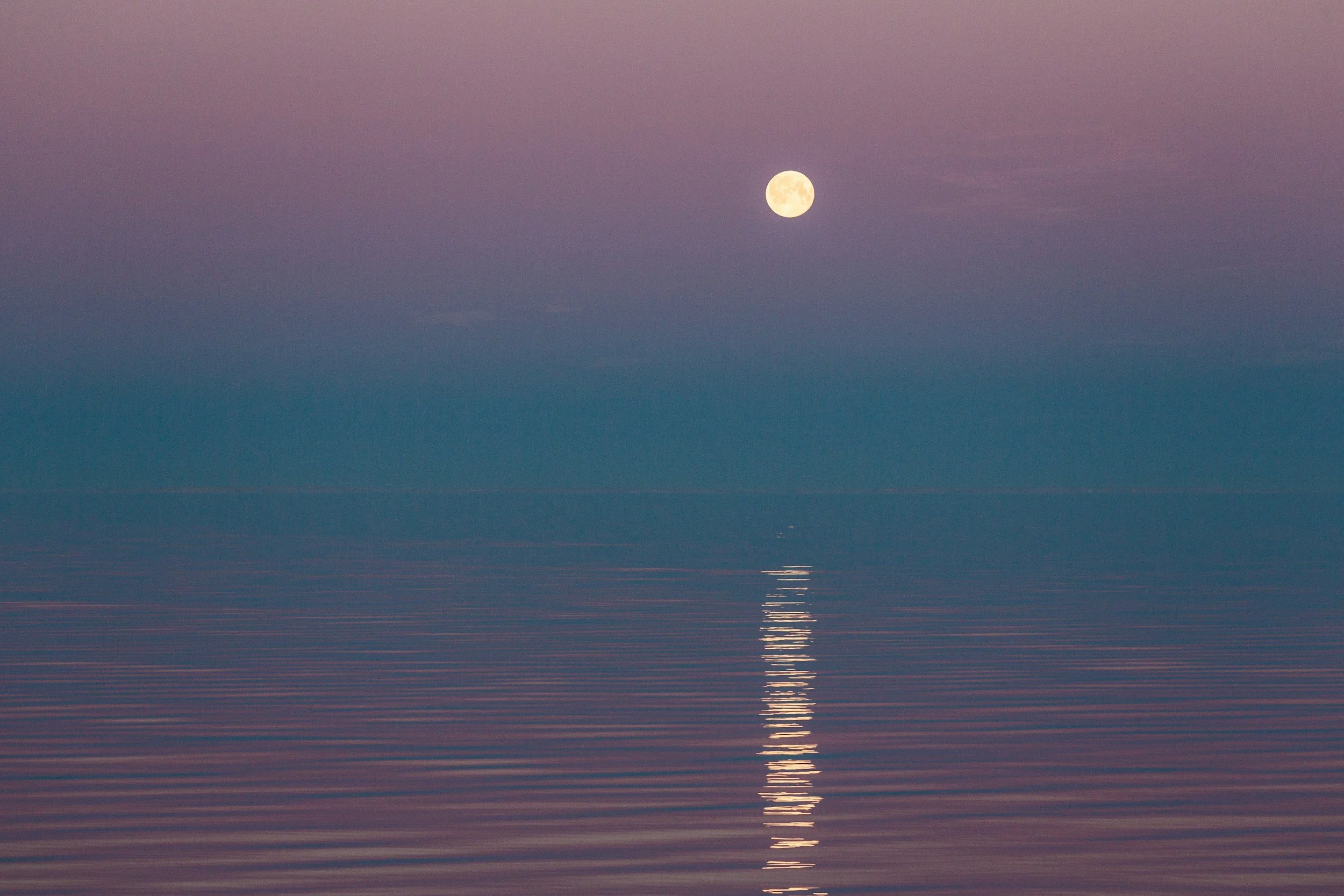 moon in the sky reflects on a blue and pink body of water.