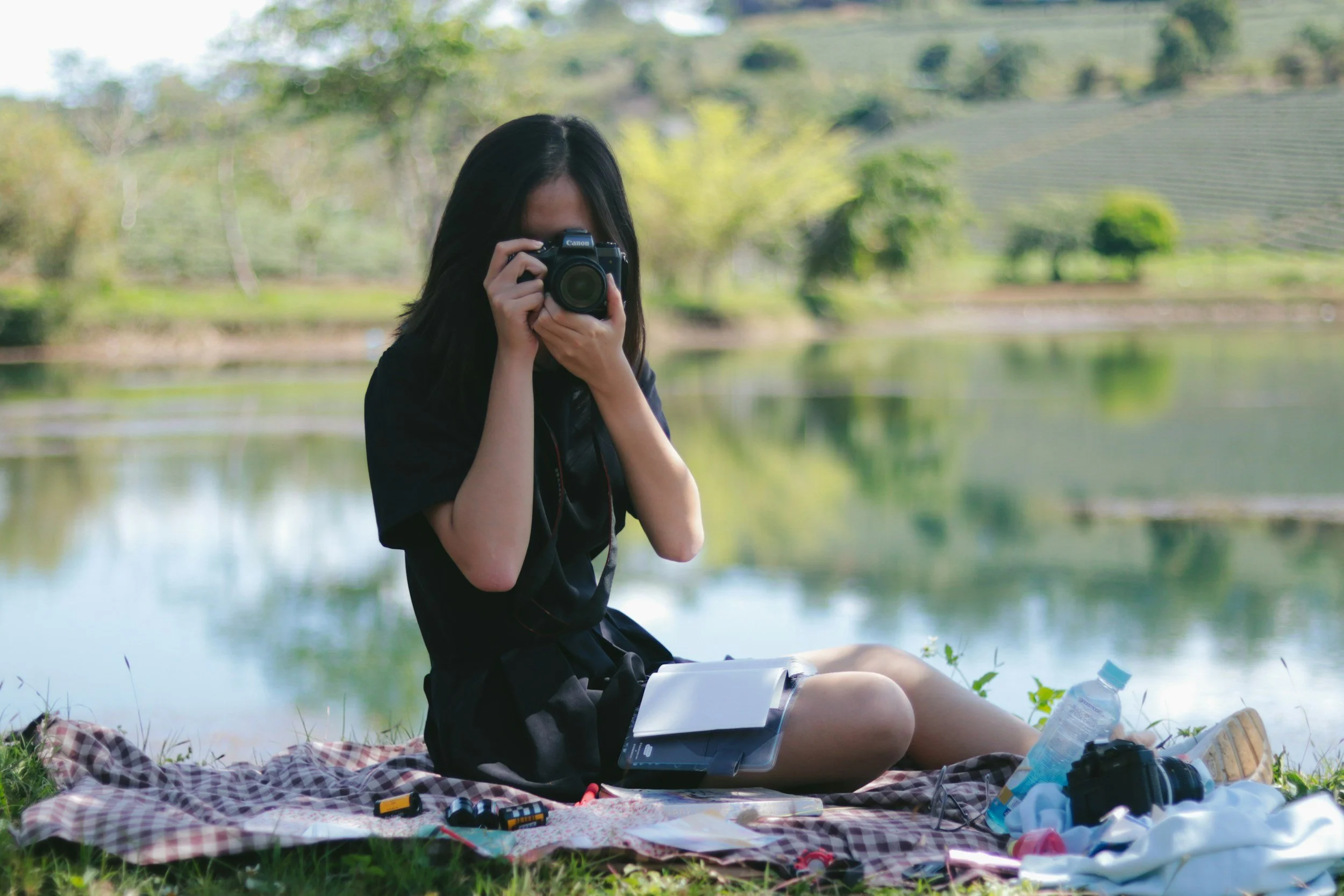a woman sits on a picnic blanket by a pond and holds a digital camera up to her eye.