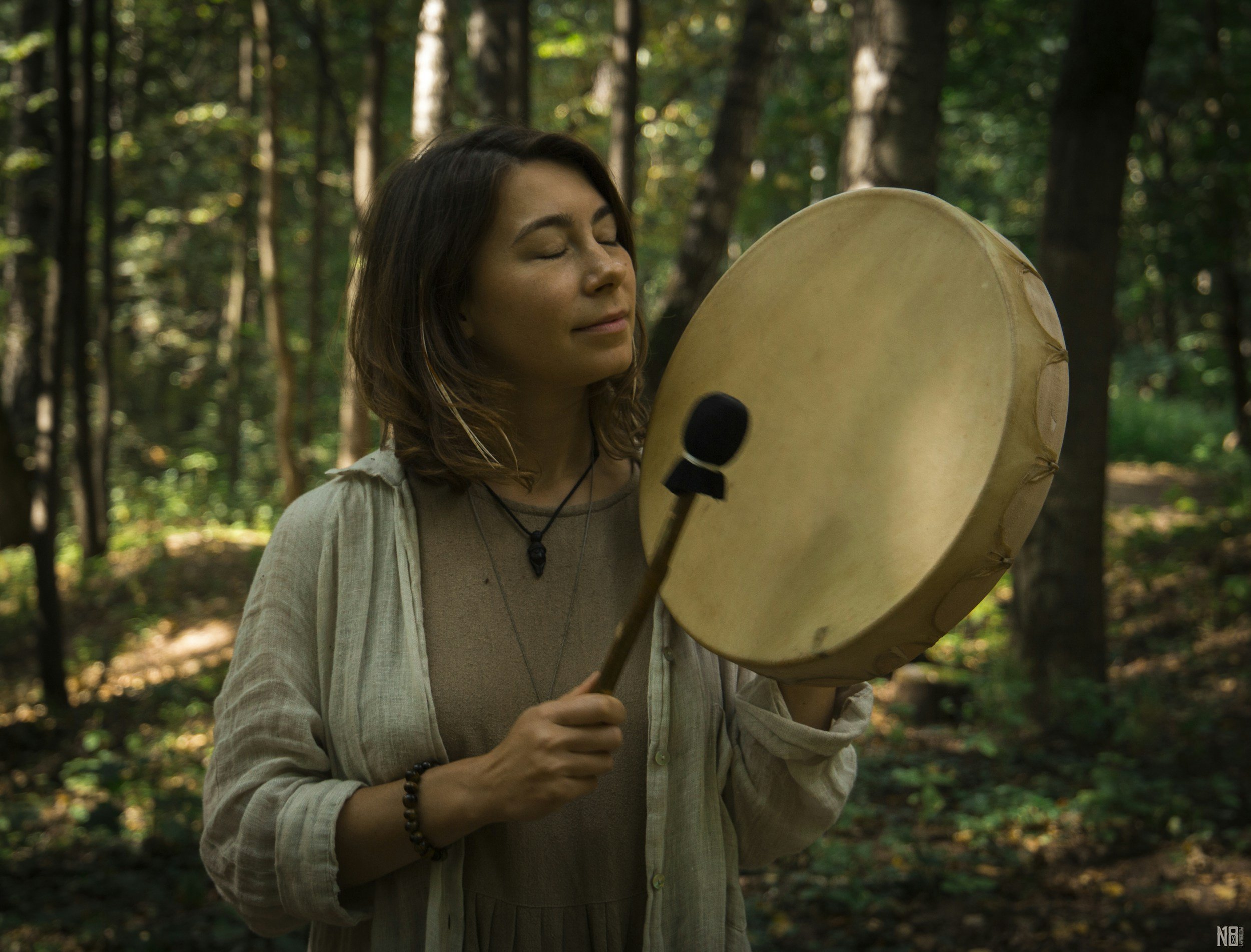 a woman stands in a forest with a simple drum and mallet.