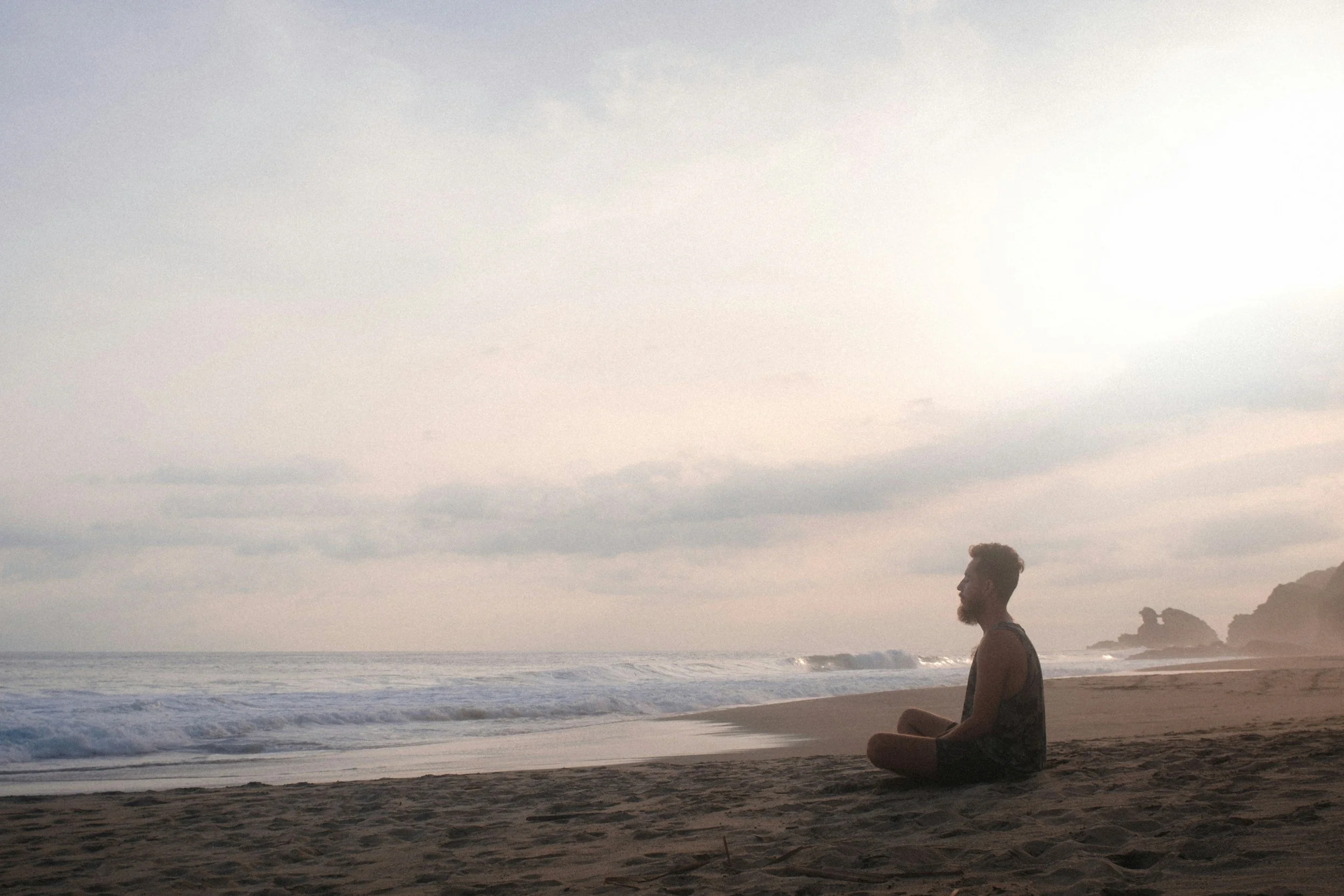 a man sits cross legged on the beach facing the water.