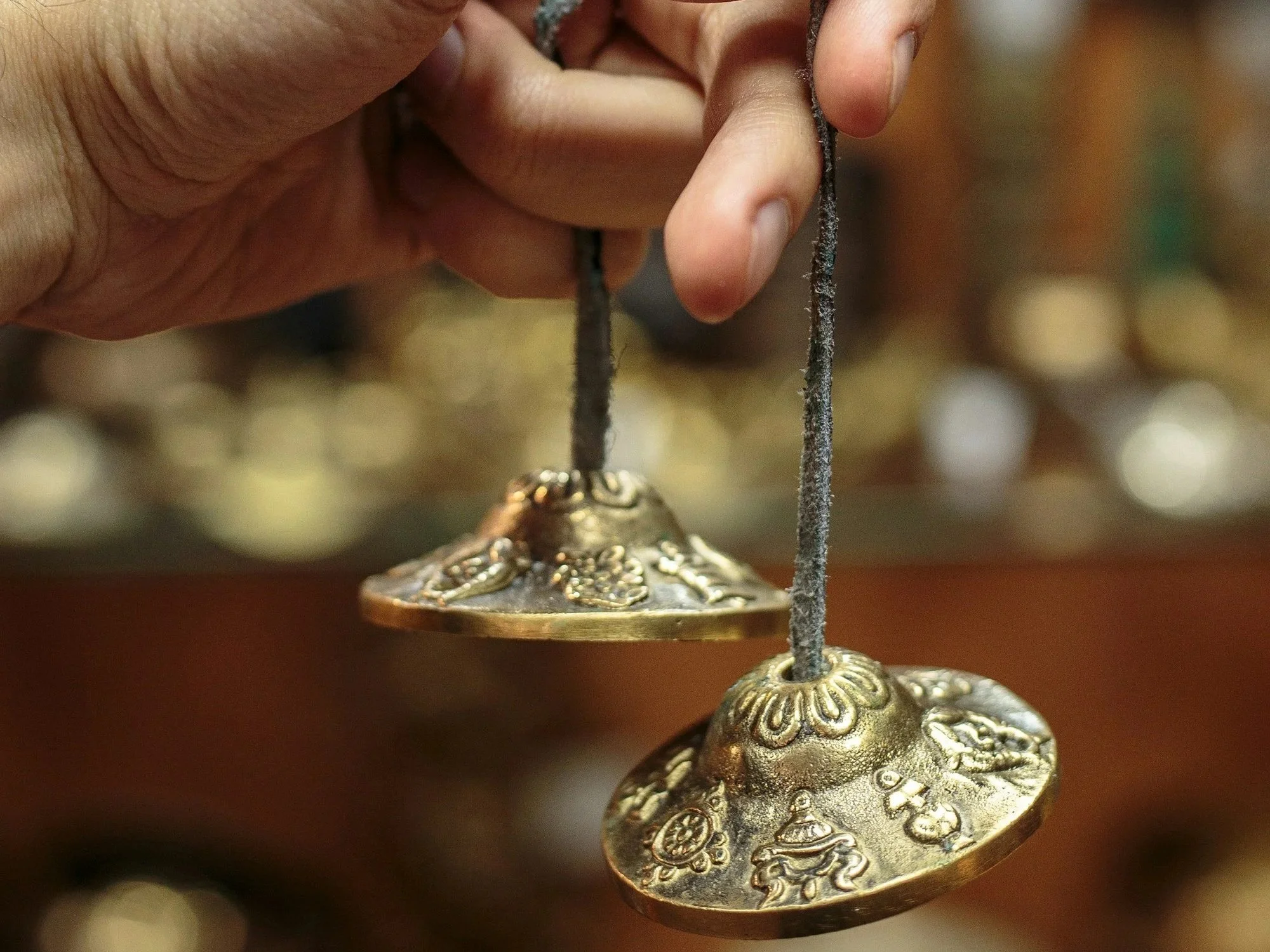 a hand holds a golden tingsha. Tingsha look like small cymbals. These ones have engraved and embossed designs.