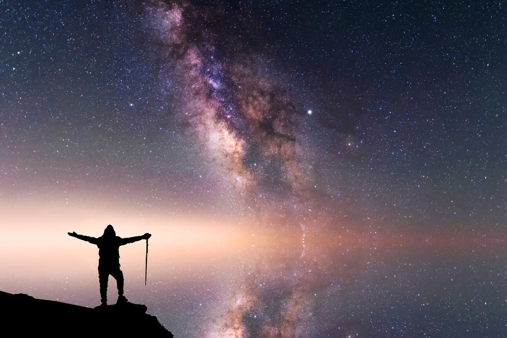 a person stands on a precipice against a background of the milkyway.