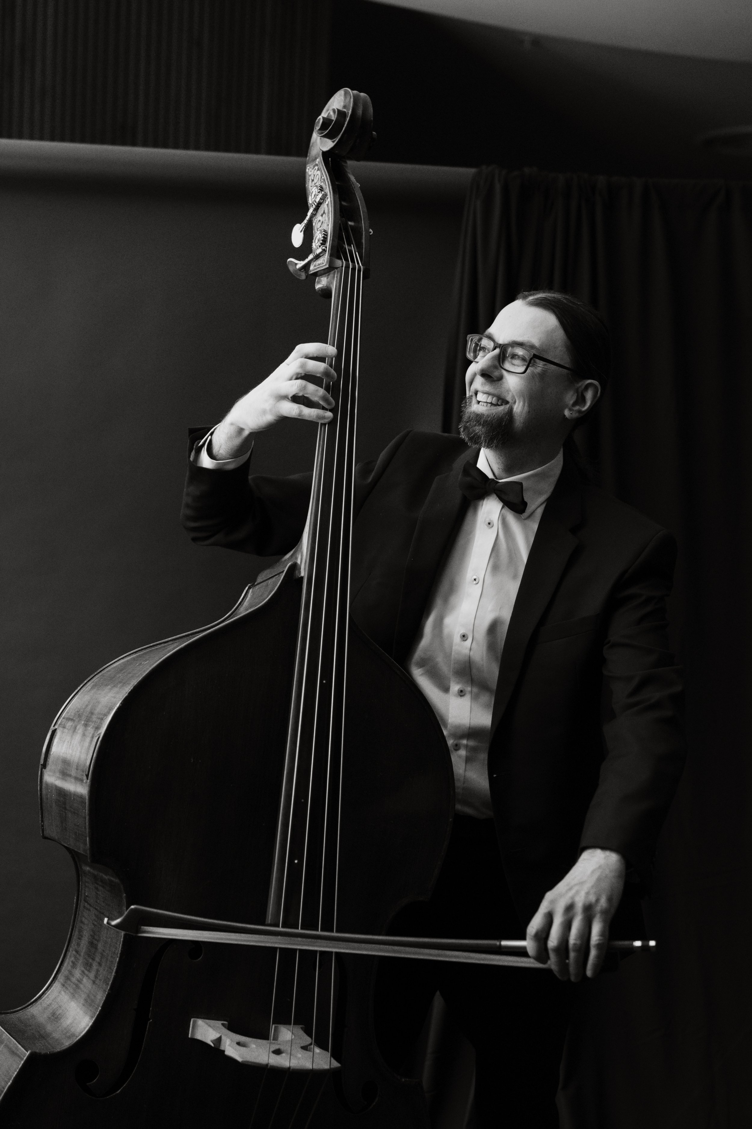 A man dressed in a tuxedo with a bow tie is smiling while playing an upright bass in a performance setting, black and white photo.