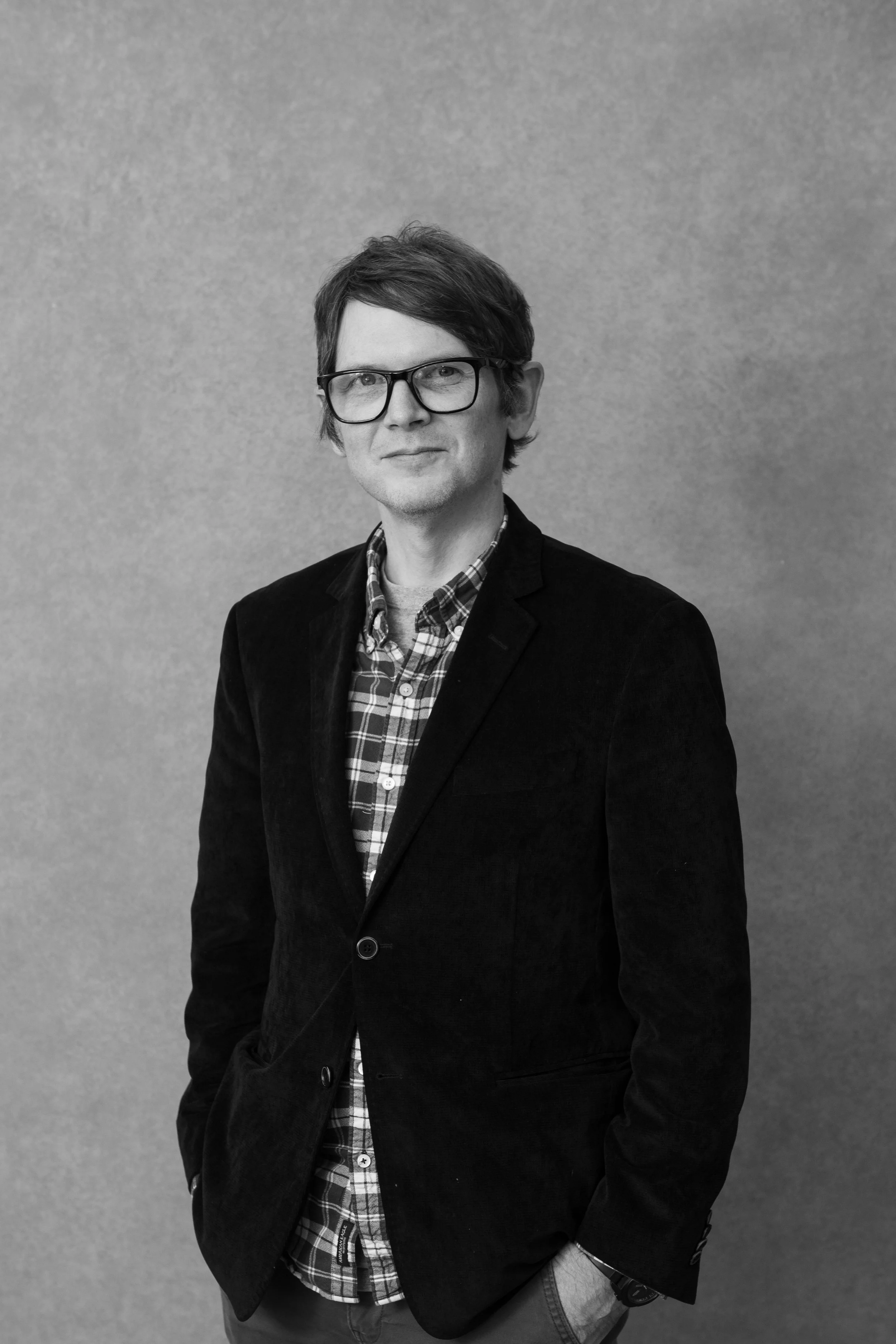 A man wearing glasses, a dark blazer, and a checkered shirt smiling against a gray background.