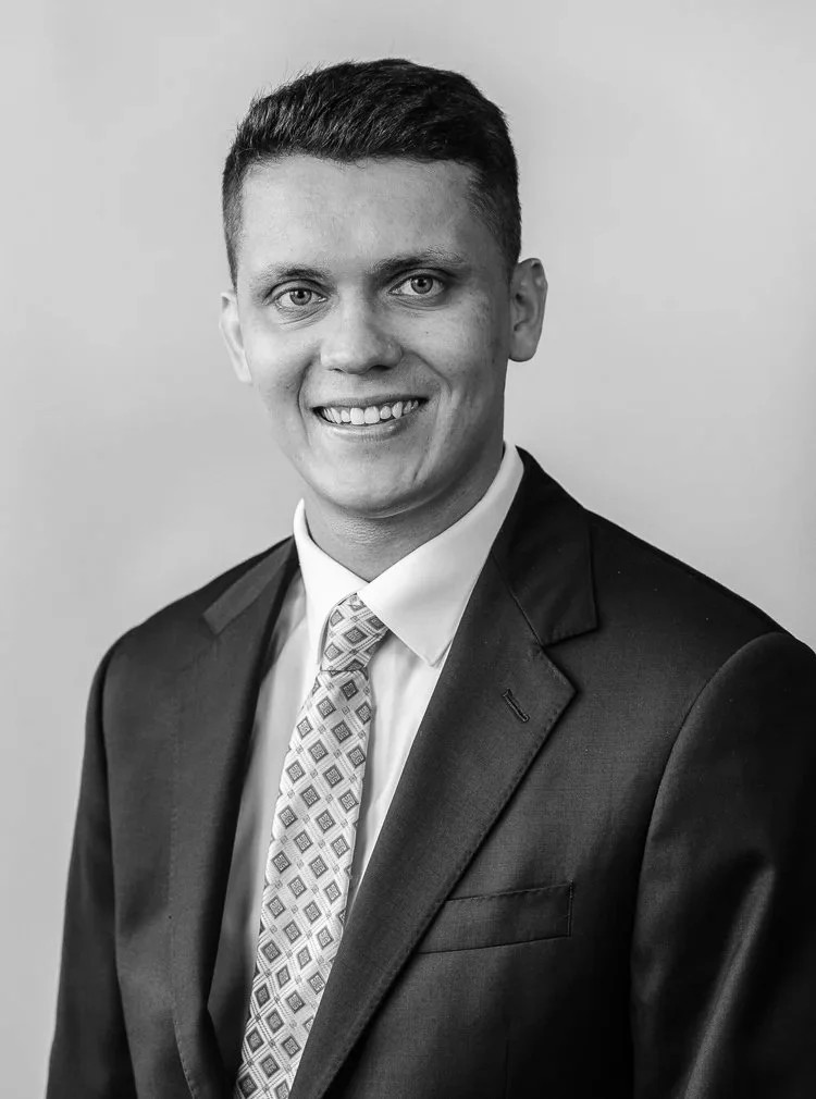 Headshot of a young man with dark hair, blue eyes, dressed in a dark blue suit, white shirt, and patterned tie, smiling against a plain light gray background.