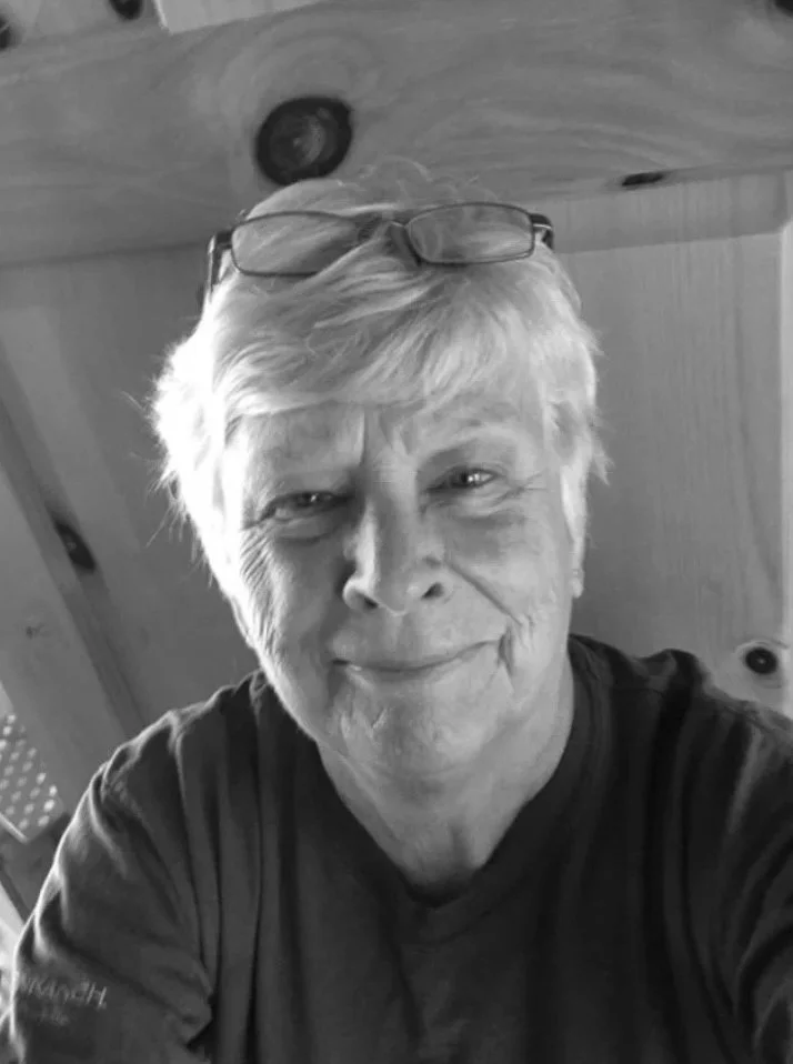 Close-up selfie of an older woman with short blonde hair, smiling, wearing glasses on her head and a dark shirt, indoors with wooden walls in the background.