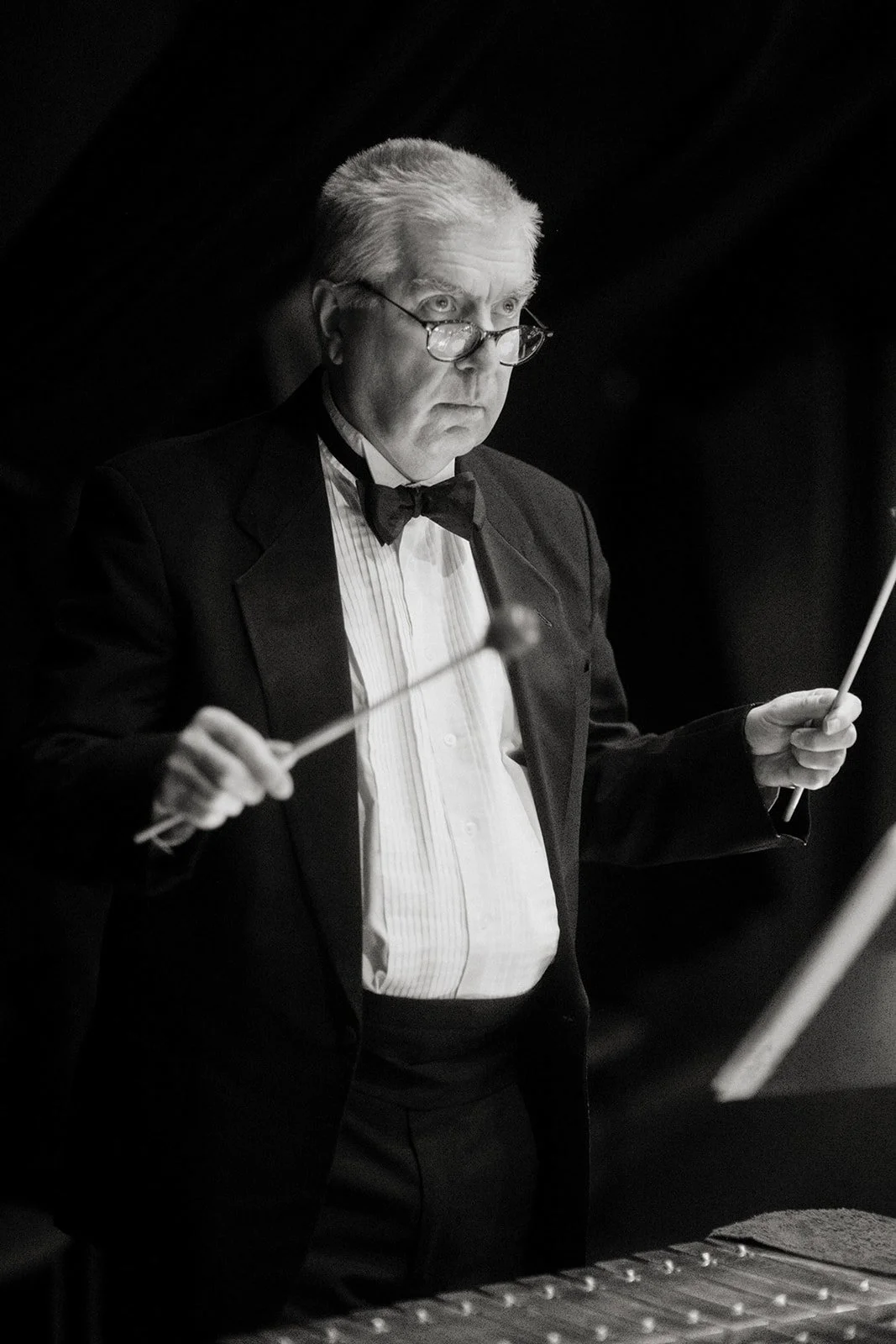 Black-and-white photo of a male orchestra conductor wearing a tuxedo, glasses, and a bowtie, holding a baton in each hand, focused while conducting.