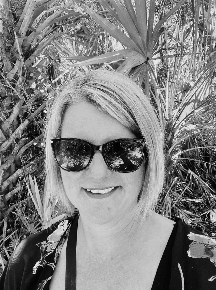 A woman with blonde hair and sunglasses smiling outdoors among green tropical plants.