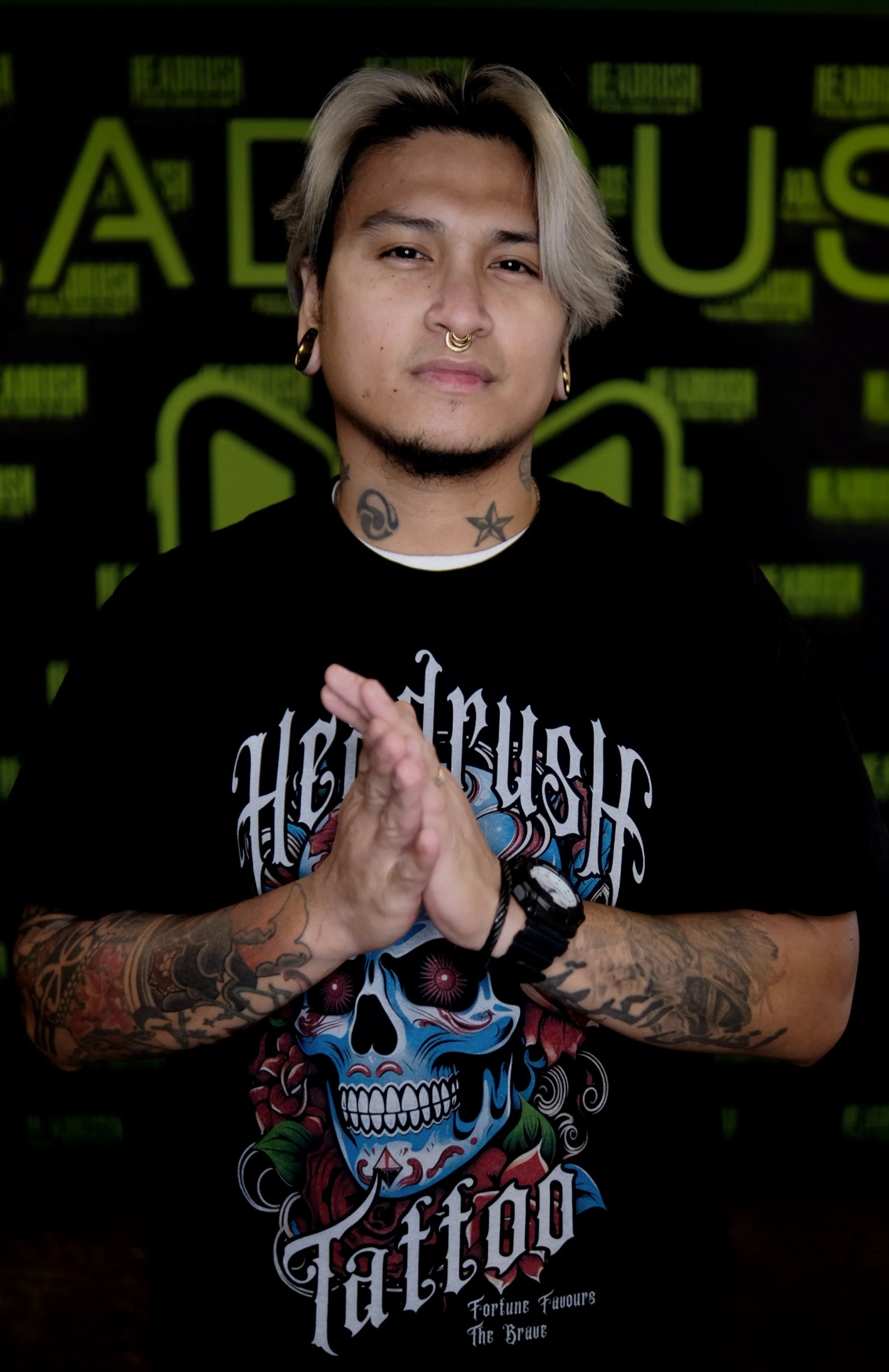 A man with blond hair and tattoos on his arms and neck, wearing a black graphic t-shirt with a colorful skull design, standing in front of a black background with green text.