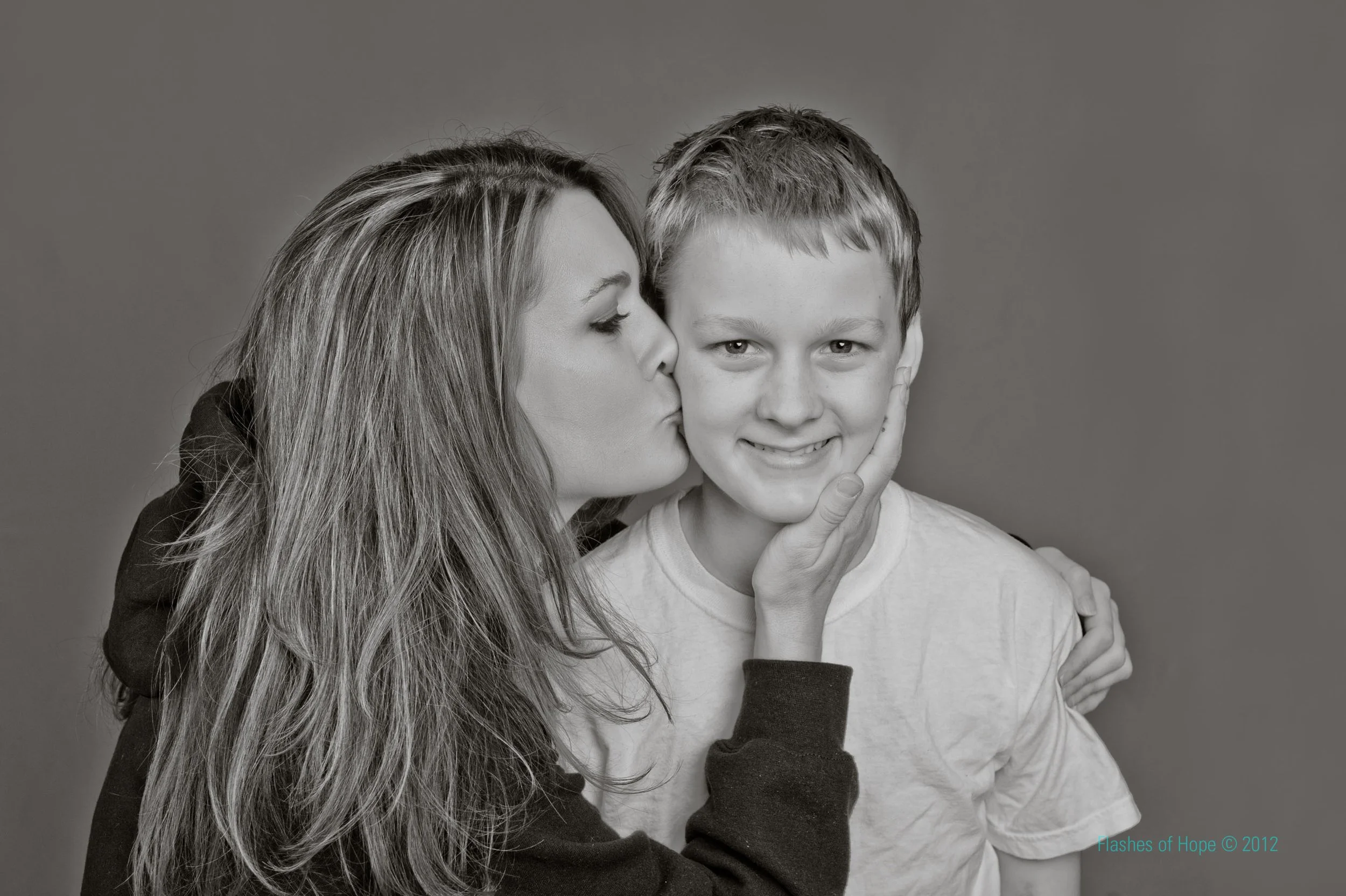 A woman with long hair is giving a gentle kiss on the cheek to a young boy with short hair. The woman has her eyes closed and appears affectionate, while the boy is smiling and touching his face with his hand. The image is in black and white.