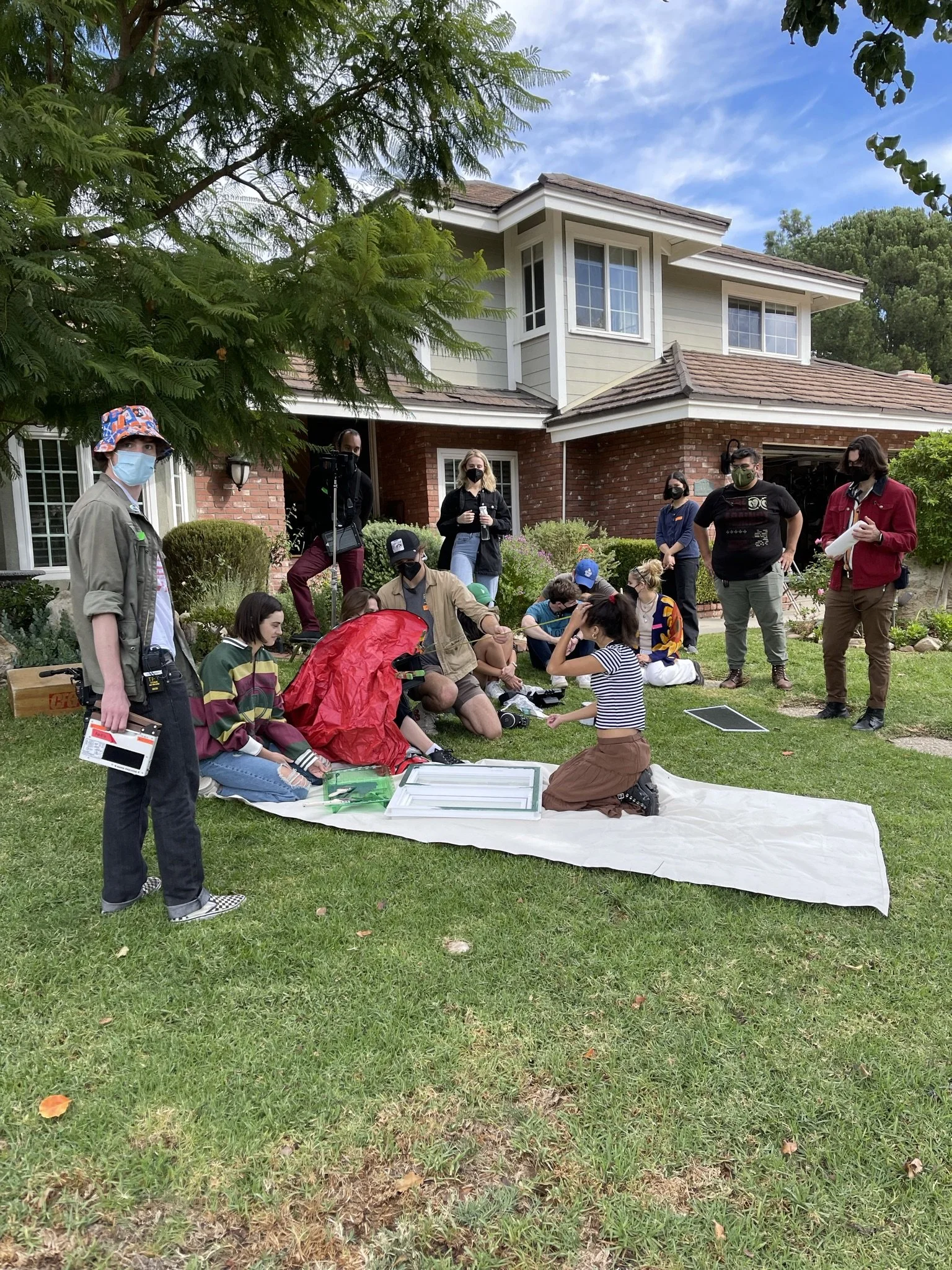 A group of people participating in an outdoor activity or workshop on the lawn of a house, with some sitting on a white mat, others standing, and all wearing masks.