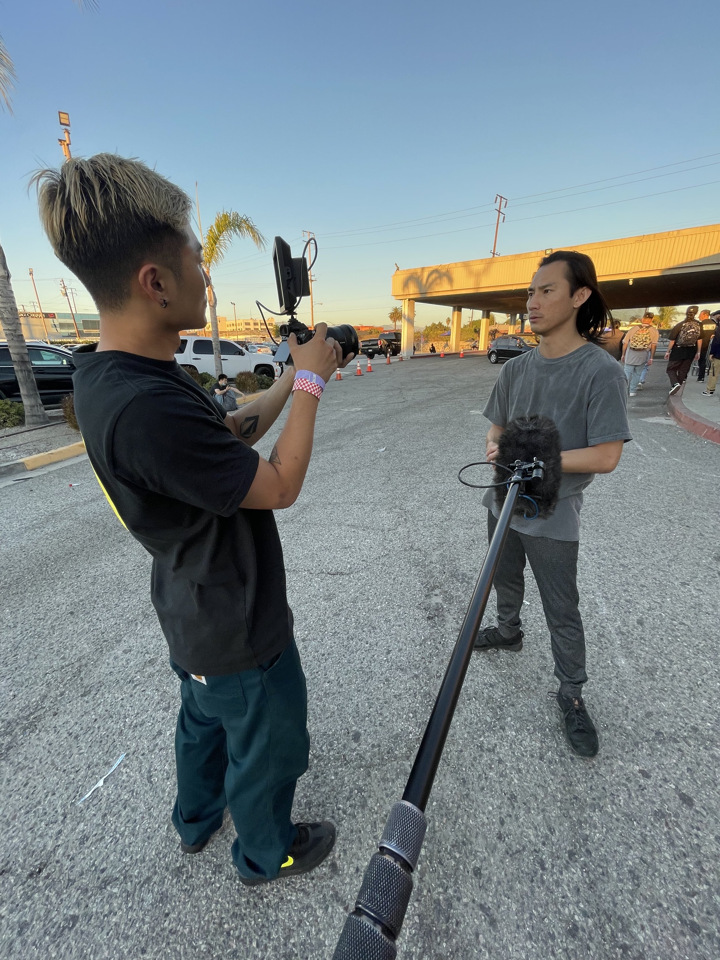 A person with long dark hair being interviewed outdoors by a person with short dyed hair, holding a camera and microphone, with cars and a building in the background during sunset.