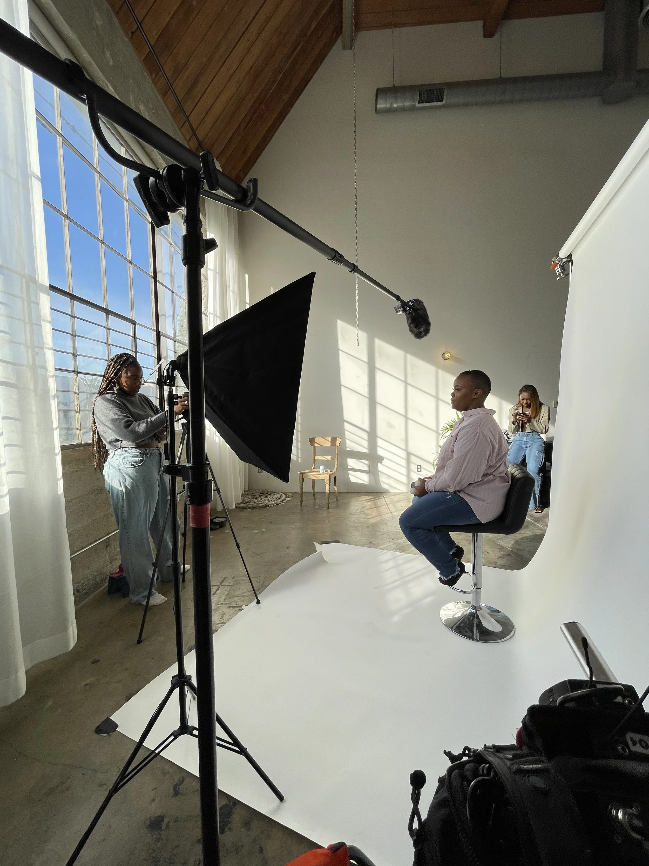 A woman sits on a stool in front of a white backdrop, being filmed for a video or photo shoot. A crew member adjusts lighting and camera equipment, with a person in the background using a phone. The studio has large windows allowing natural light.