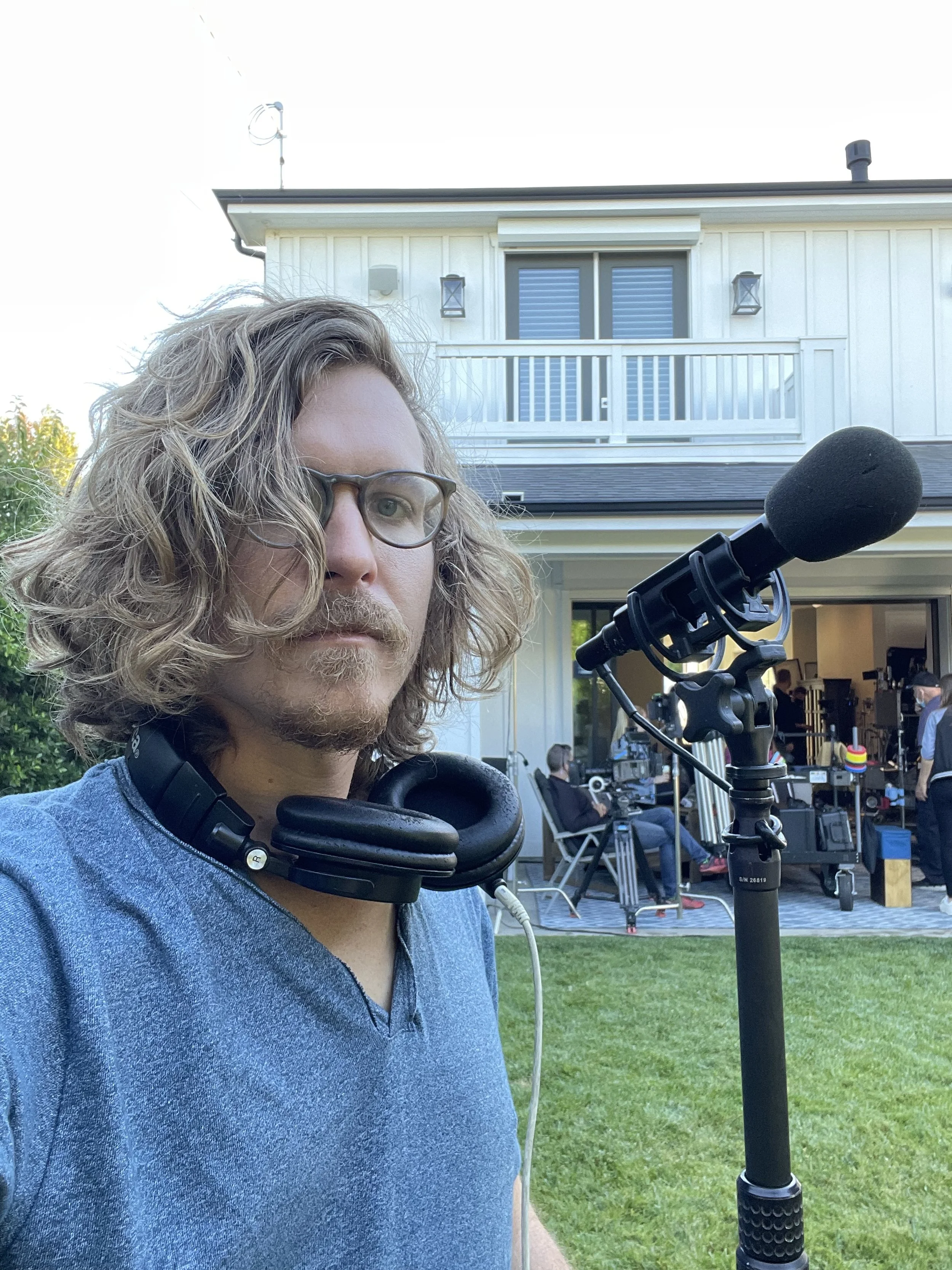 A man with long wavy hair, glasses, and a beard standing outdoors in front of a house, with a sound microphone in front of him and film crew working in the background.