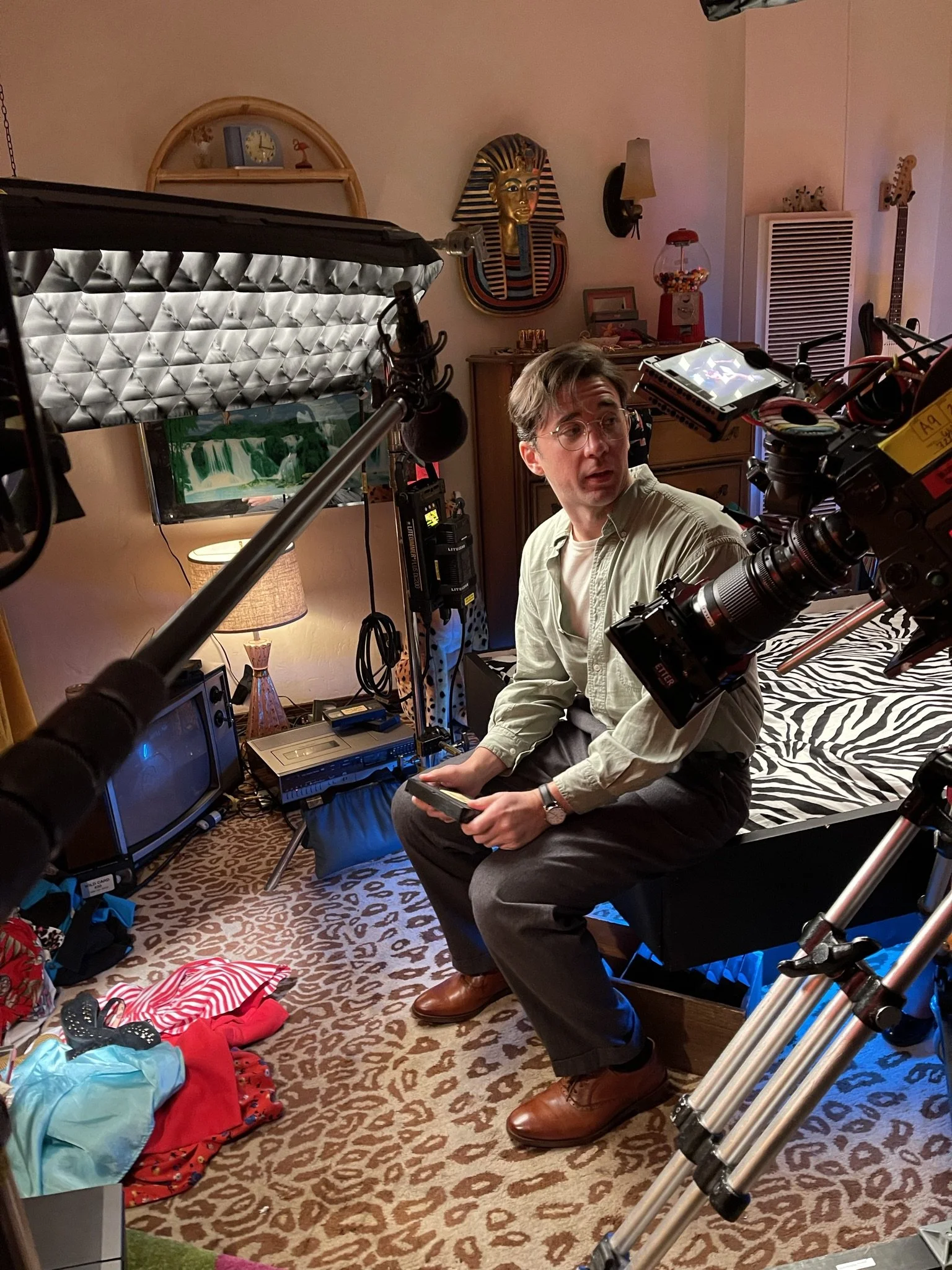 A man with glasses, wearing a light-colored button-up shirt, sitting on a bench in a cluttered room with filming equipment around him, including lights and cameras, with colorful household decor and furniture visible.