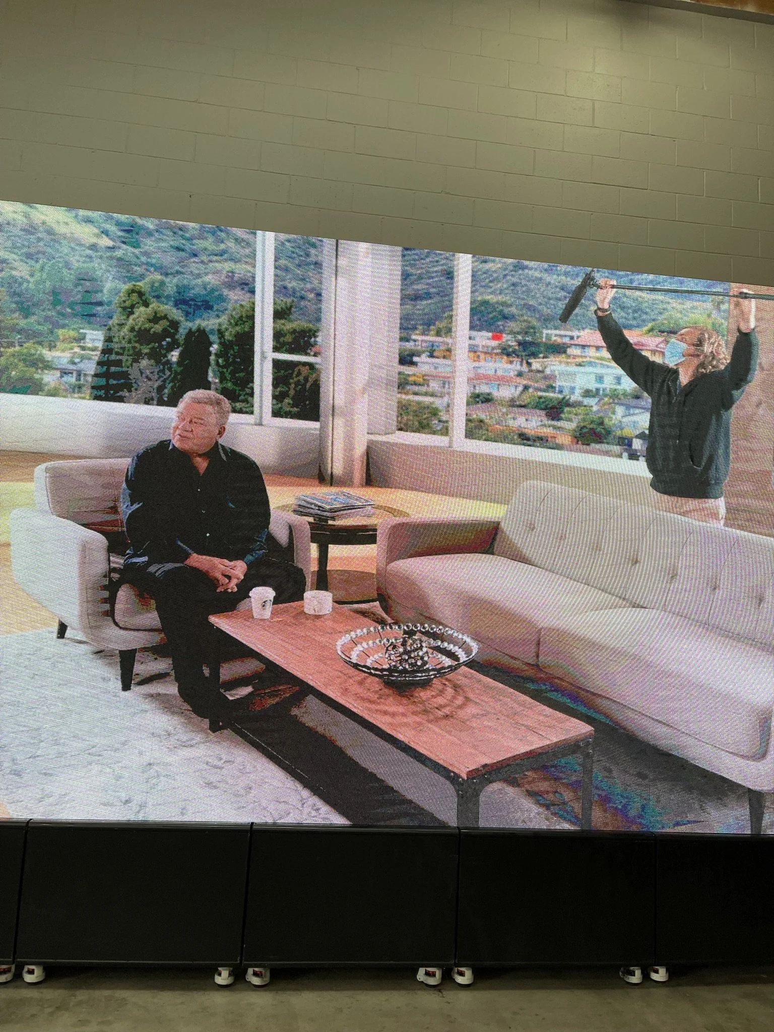 A woman wearing a face mask and holding a microphone overhead stands in a modern, well-lit room with large windows showing city and mountain views. A man is seated on a white sofa, facing slightly away, with a small table holding cups and magazines i