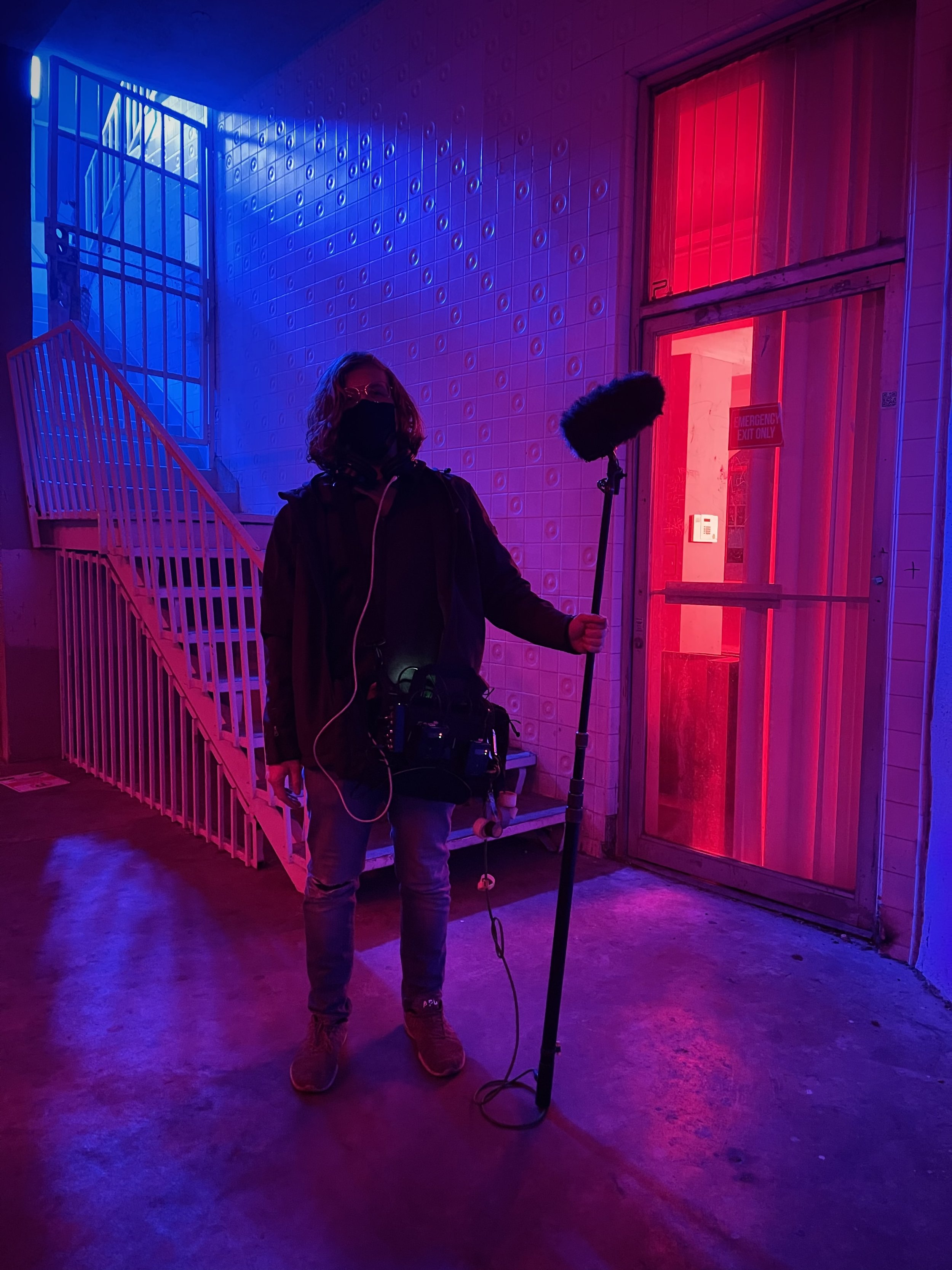 A person wearing a black jacket, jeans, and a face mask holding a boom microphone inside a room with vibrant pink and blue lighting, next to a pink door with glass.