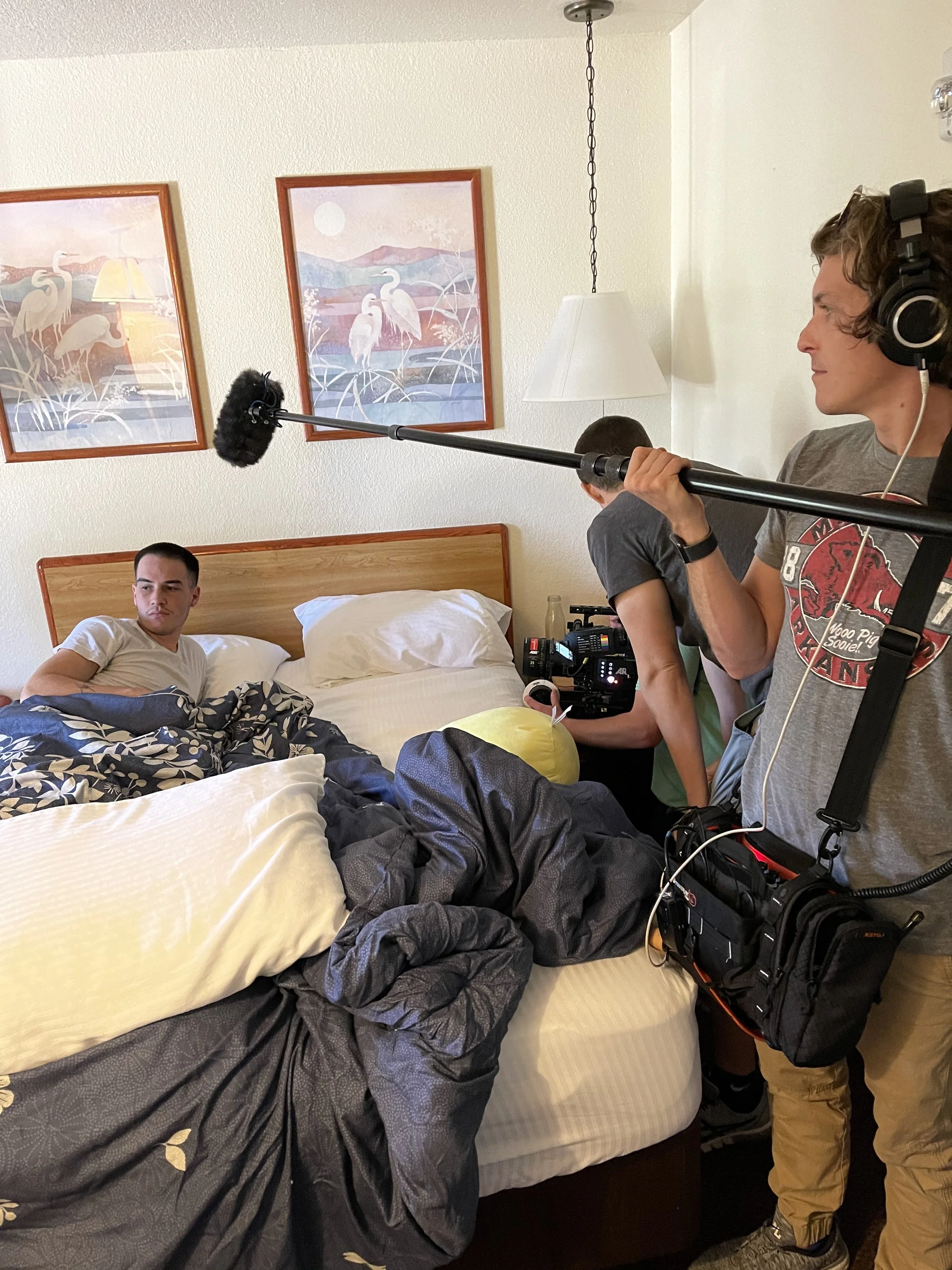 A film crew is filming a scene in a bedroom with a young man lying in bed. The young man is looking at the crew, who are setting up equipment including a camera and a microphone.
