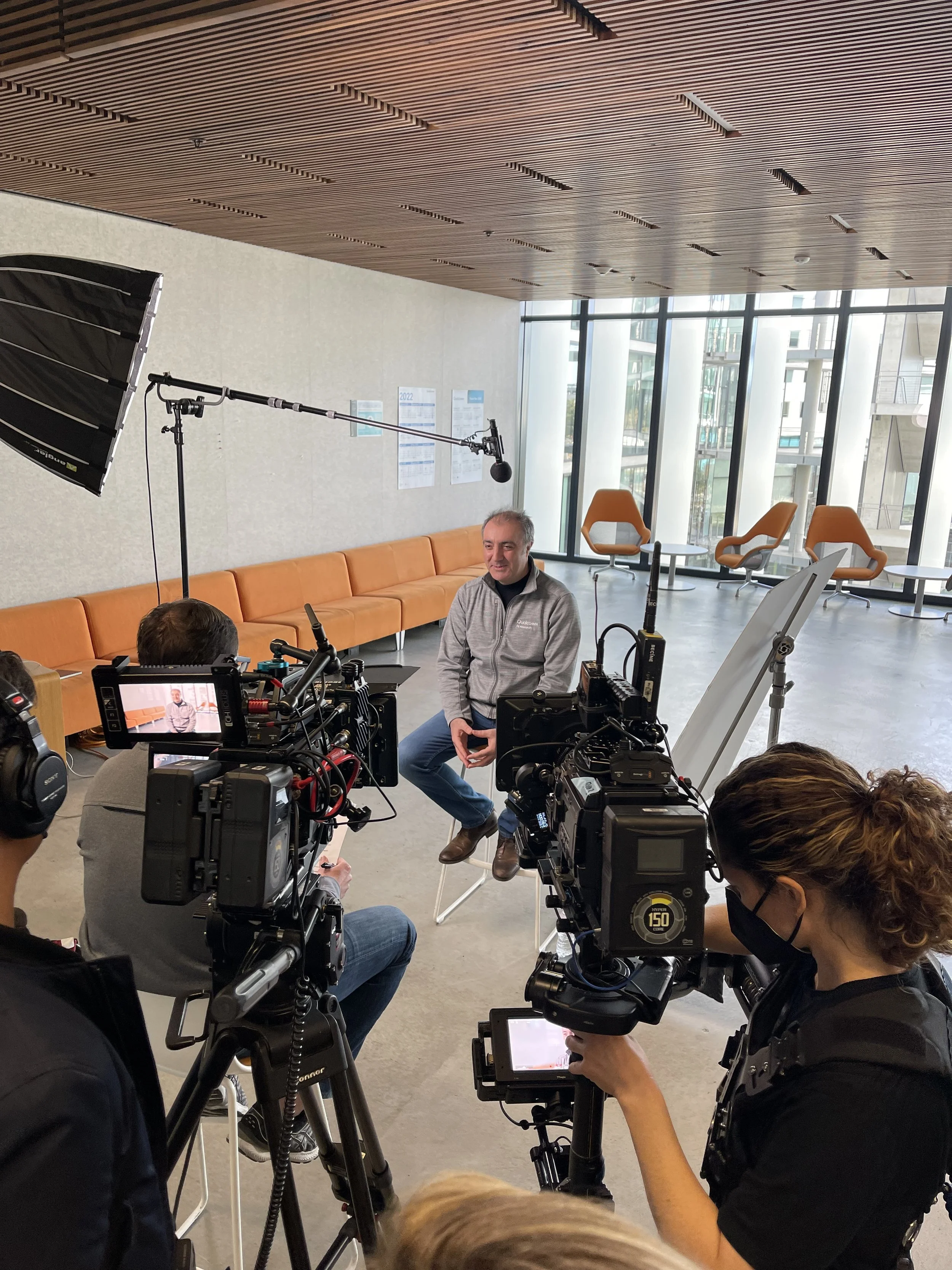 A man in a gray jacket sitting on a stool being filmed by a camera crew in a modern room with large windows, orange chairs, and wood ceiling.