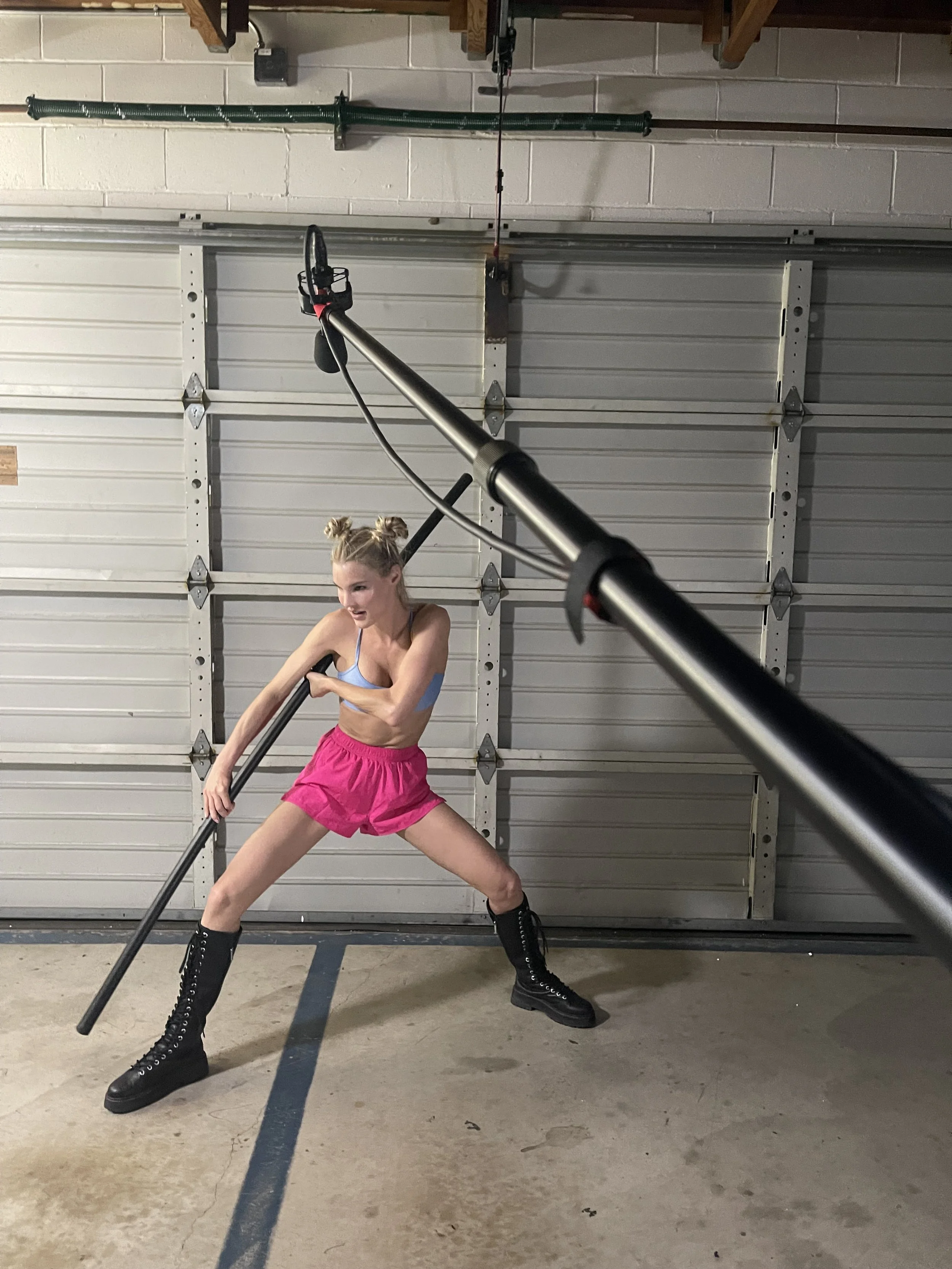 A woman dressed in a blue top, pink shorts, and black combat boots is practicing pole dancing in a garage, holding a pole with both hands while positioned with her legs spread apart.