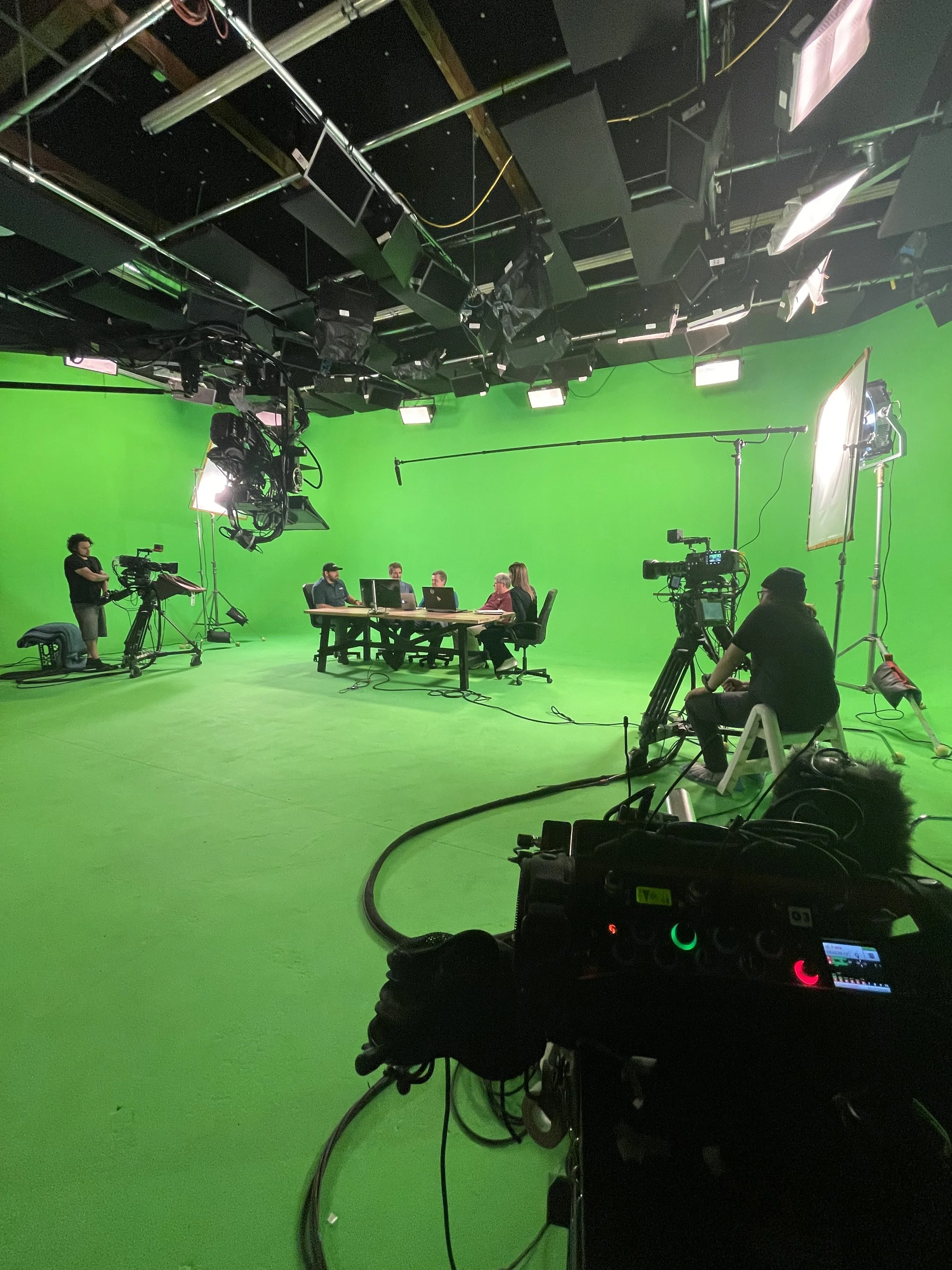 Green screen studio with a table of five people, cameras, lighting, and crew preparing for a video shoot or broadcast.