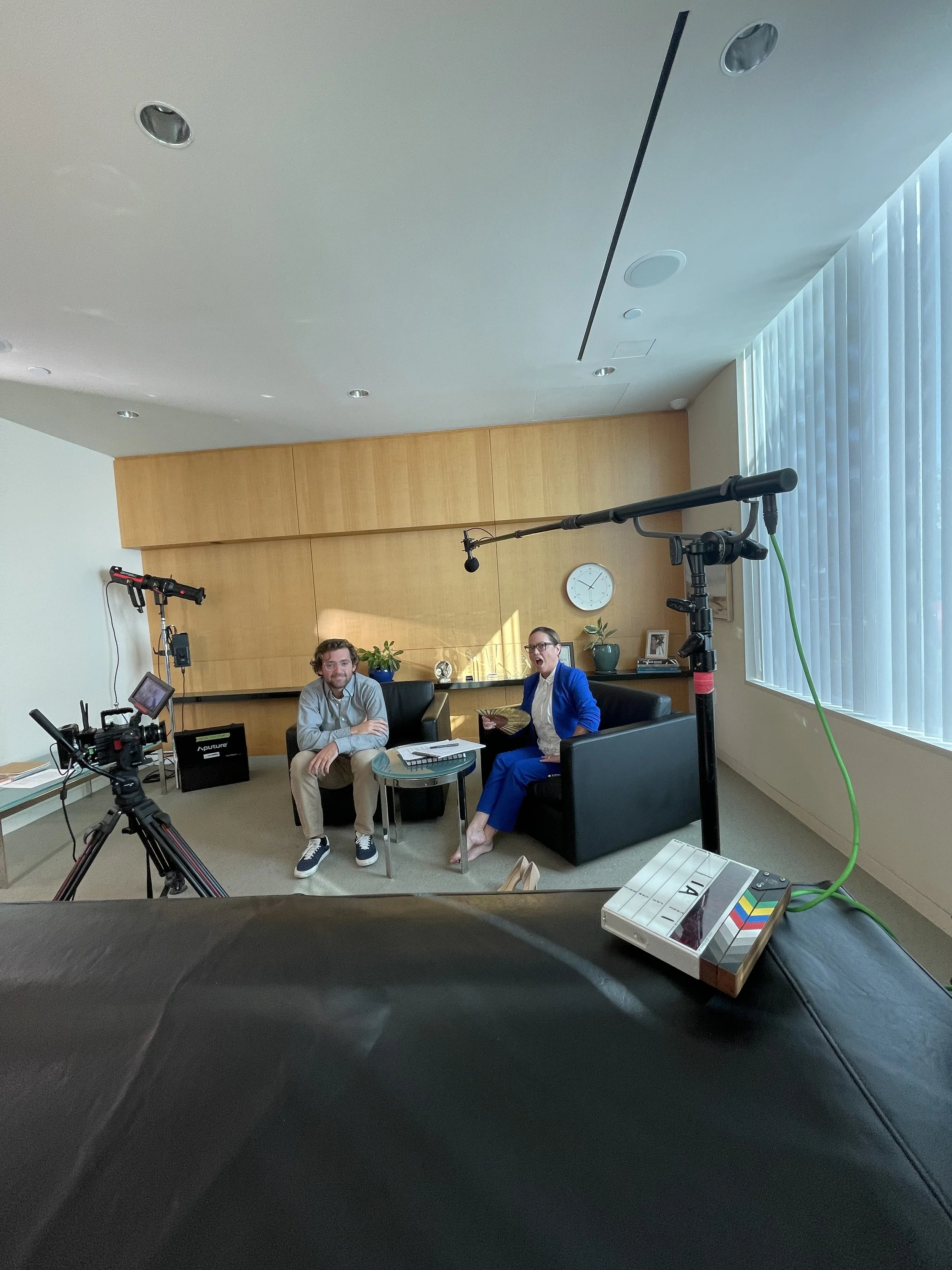 Two people being filmed in an office setting with cameras and lighting equipment, sitting on black couches with a glass coffee table between them, one dressed casually and the other in a blue suit, smiling and engaging in conversation.