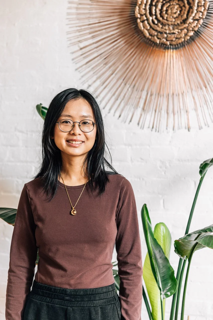 Portrait of a young Asian woman with long black hair, wearing glasses and a black top, smiling against a plain gray background.