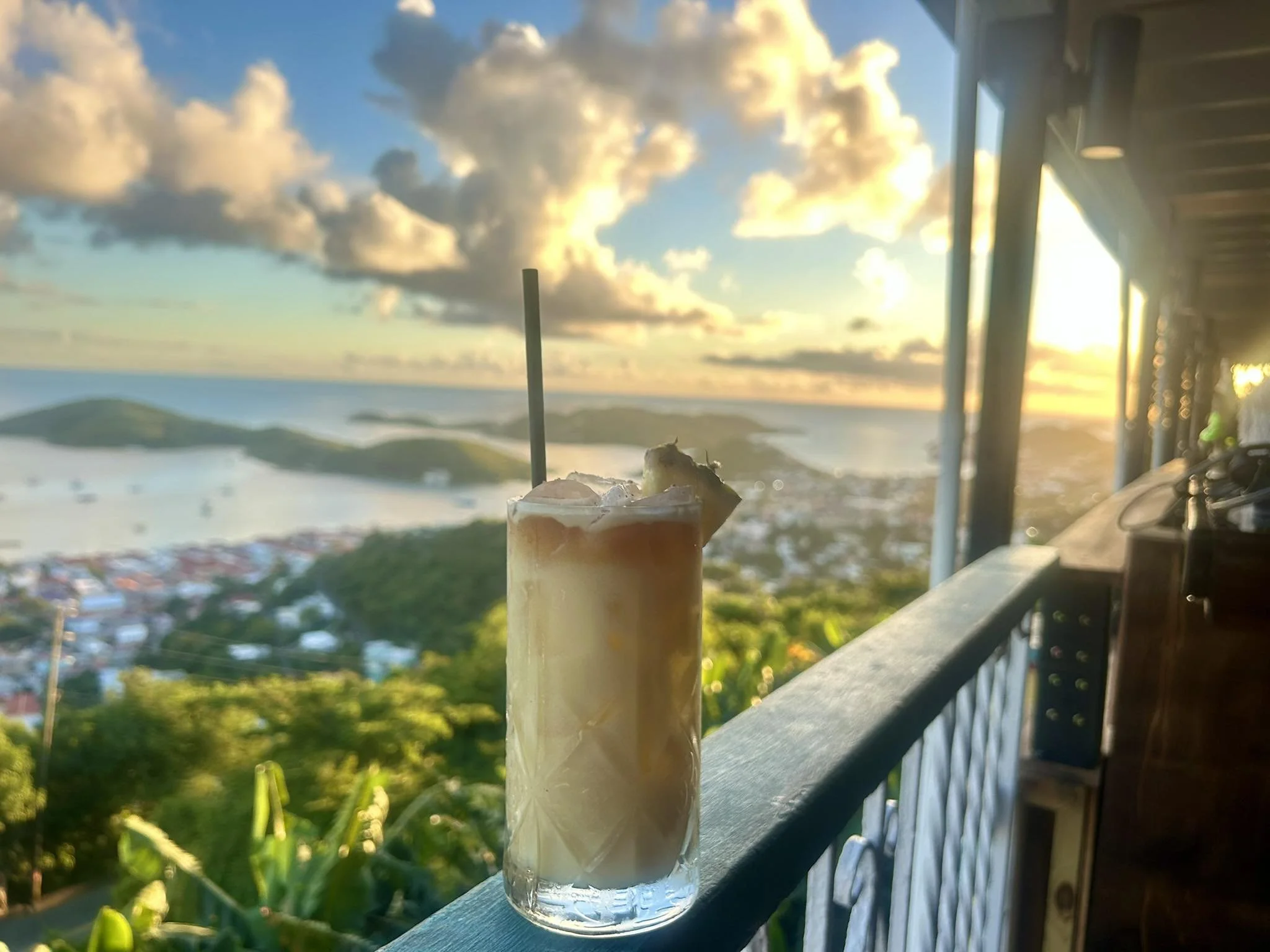 A tropical drink with ice and a straw on a balcony, overlooking a sunset over a landscape with hills and water.