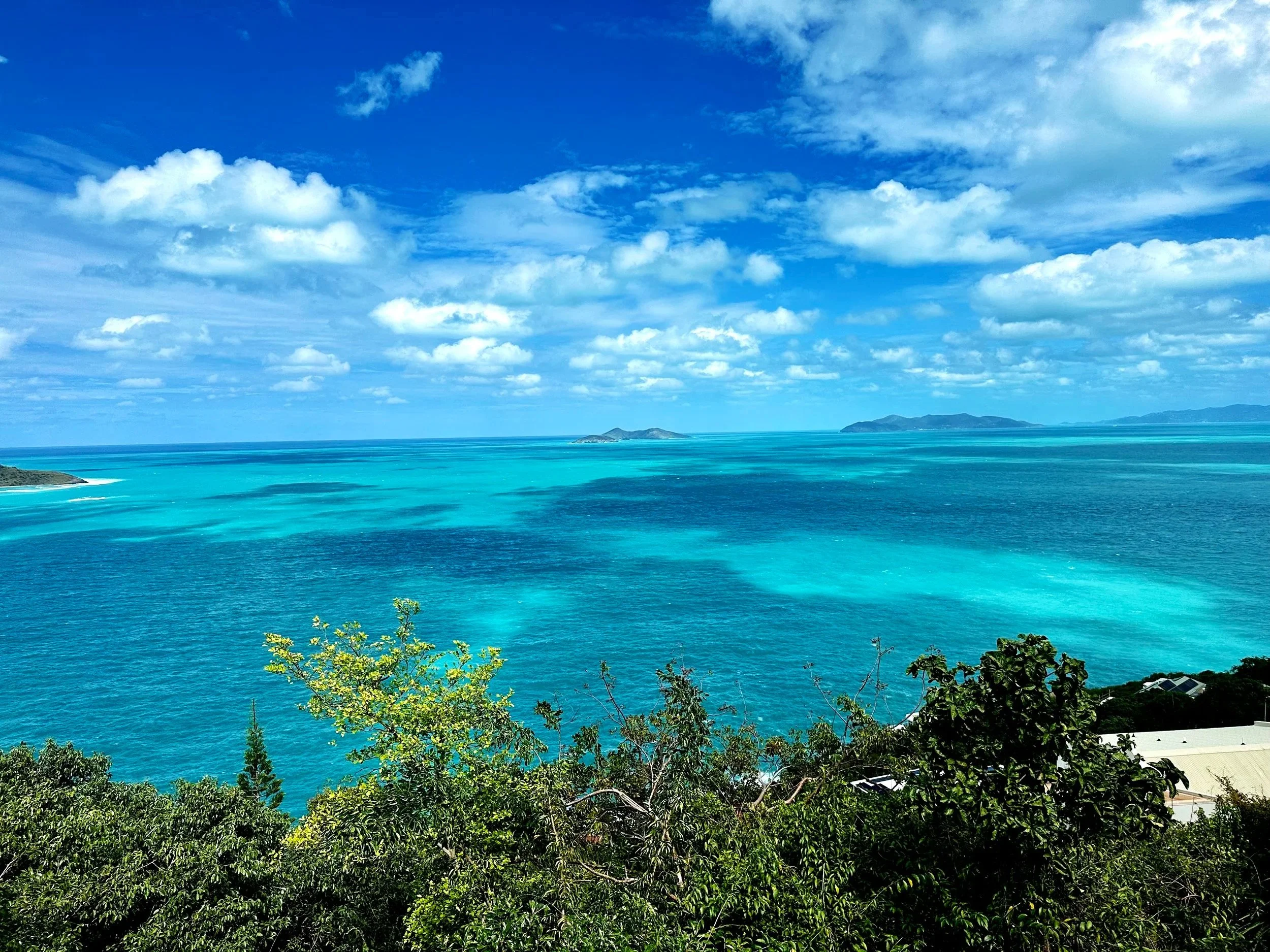 A scenic view of a turquoise ocean with lush green foliage in the foreground, a partly cloudy blue sky, and distant islands on the horizon.
