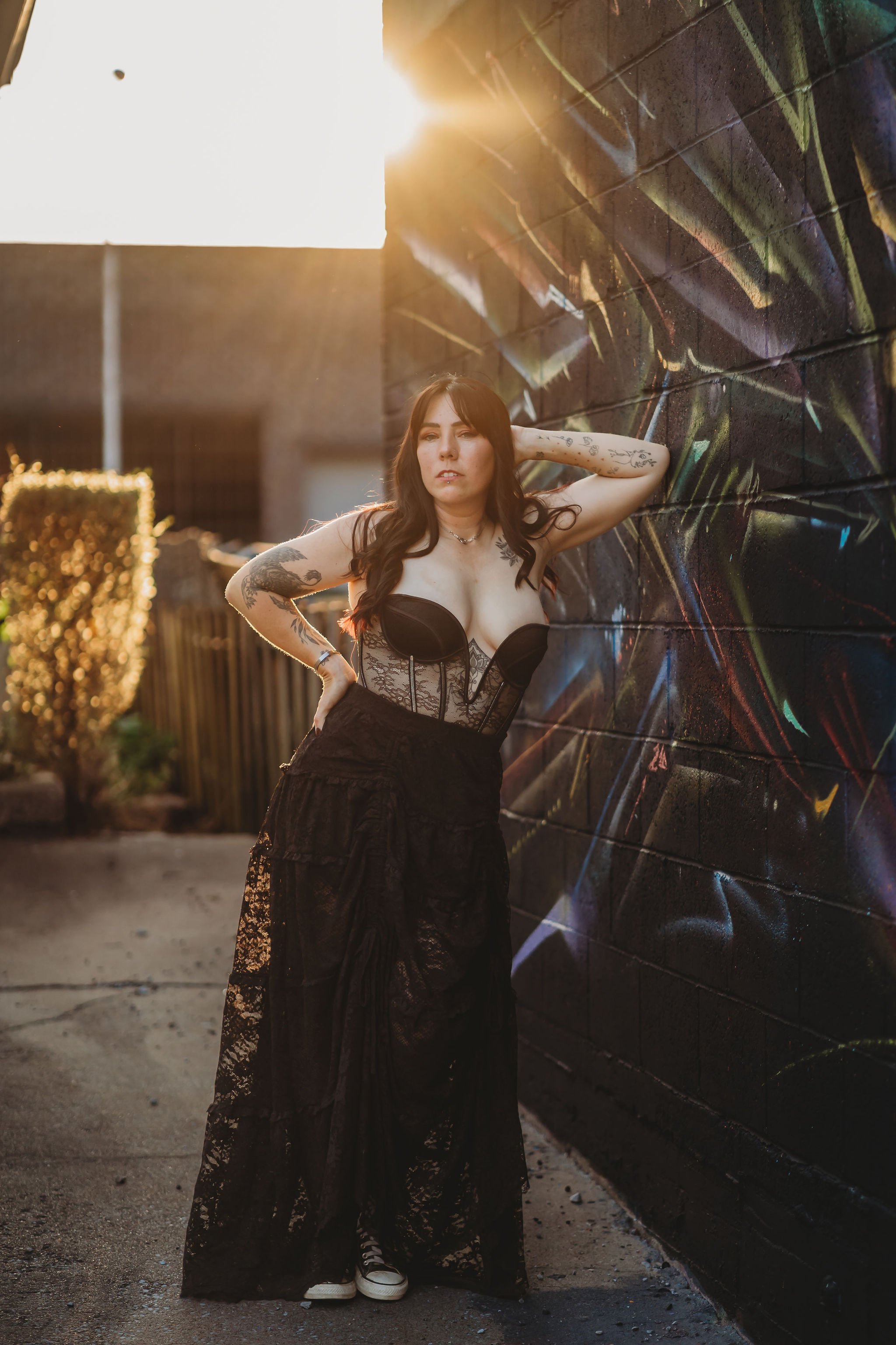 A woman with dark hair and tattoos wearing a black corset and black lace skirt stands against a graffitied wall in sunlight, with her left arm behind her head and her right hand on her hip, wearing sneakers.