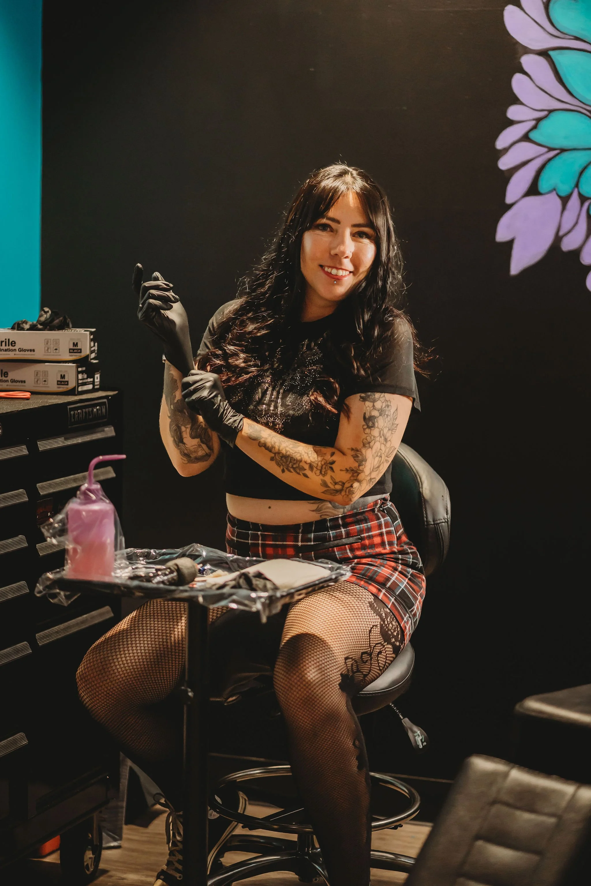 A woman with long dark hair and tattoos on her arms and thigh is sitting on a black rolling stool in a tattoo shop. She is wearing fishnet stockings, a black crop top, and red plaid skirt. She is smiling and holding black rubber gloves. The backgroun