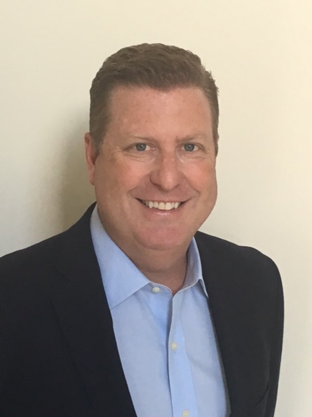 A man with short brown hair wearing a dark suit jacket and a light blue dress shirt, smiling, standing against a plain light-colored wall.